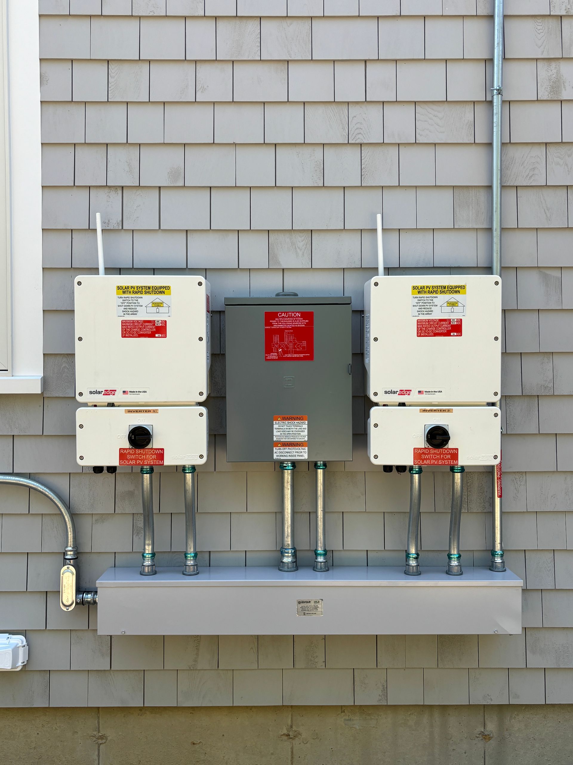 Solar power equipment mounted on gray shingled wall: inverters, combiner box, and conduit.