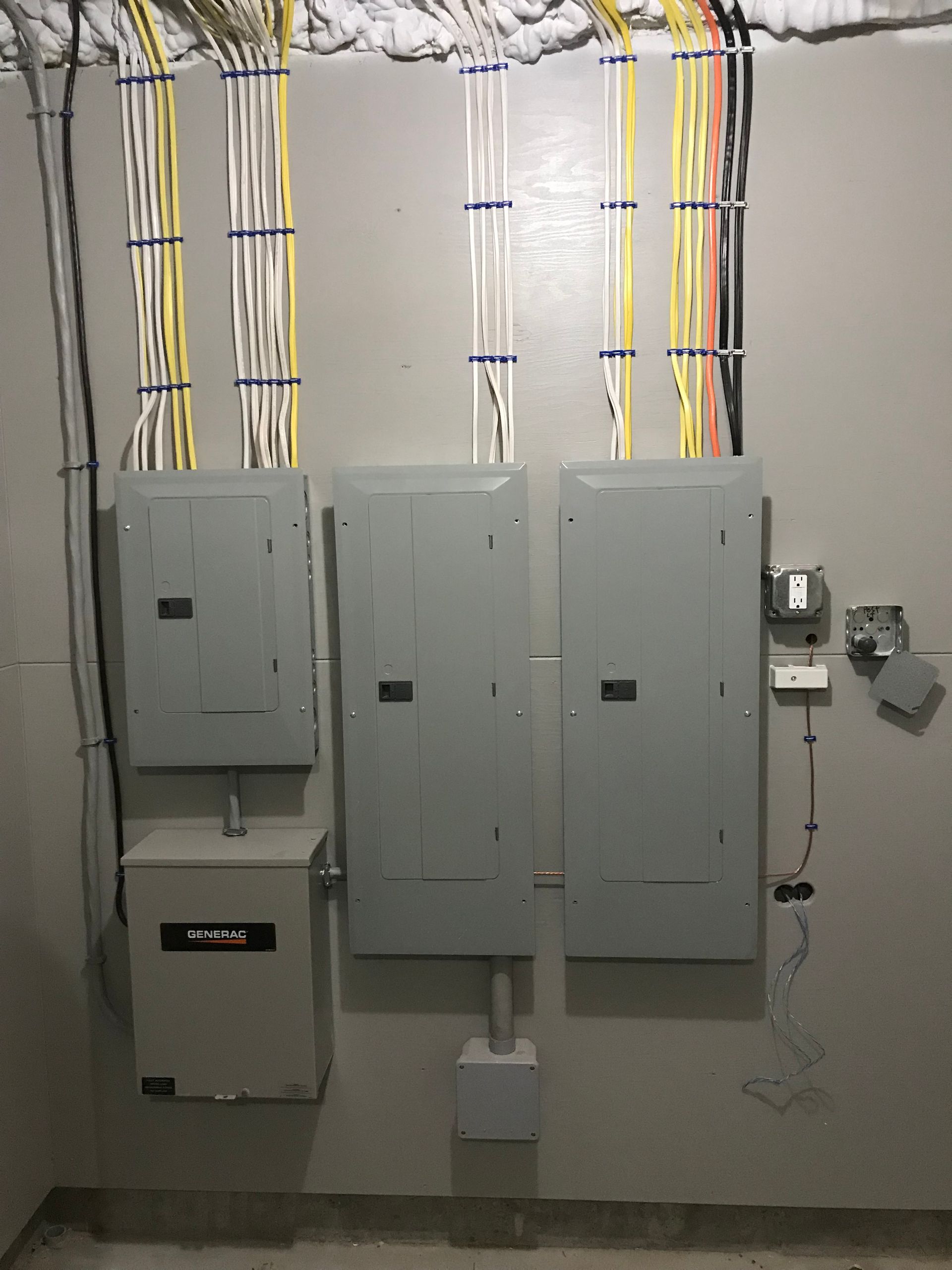 Electrical panels and wiring on a gray wall. Three gray breaker boxes and a small safe, with cables running to the ceiling.