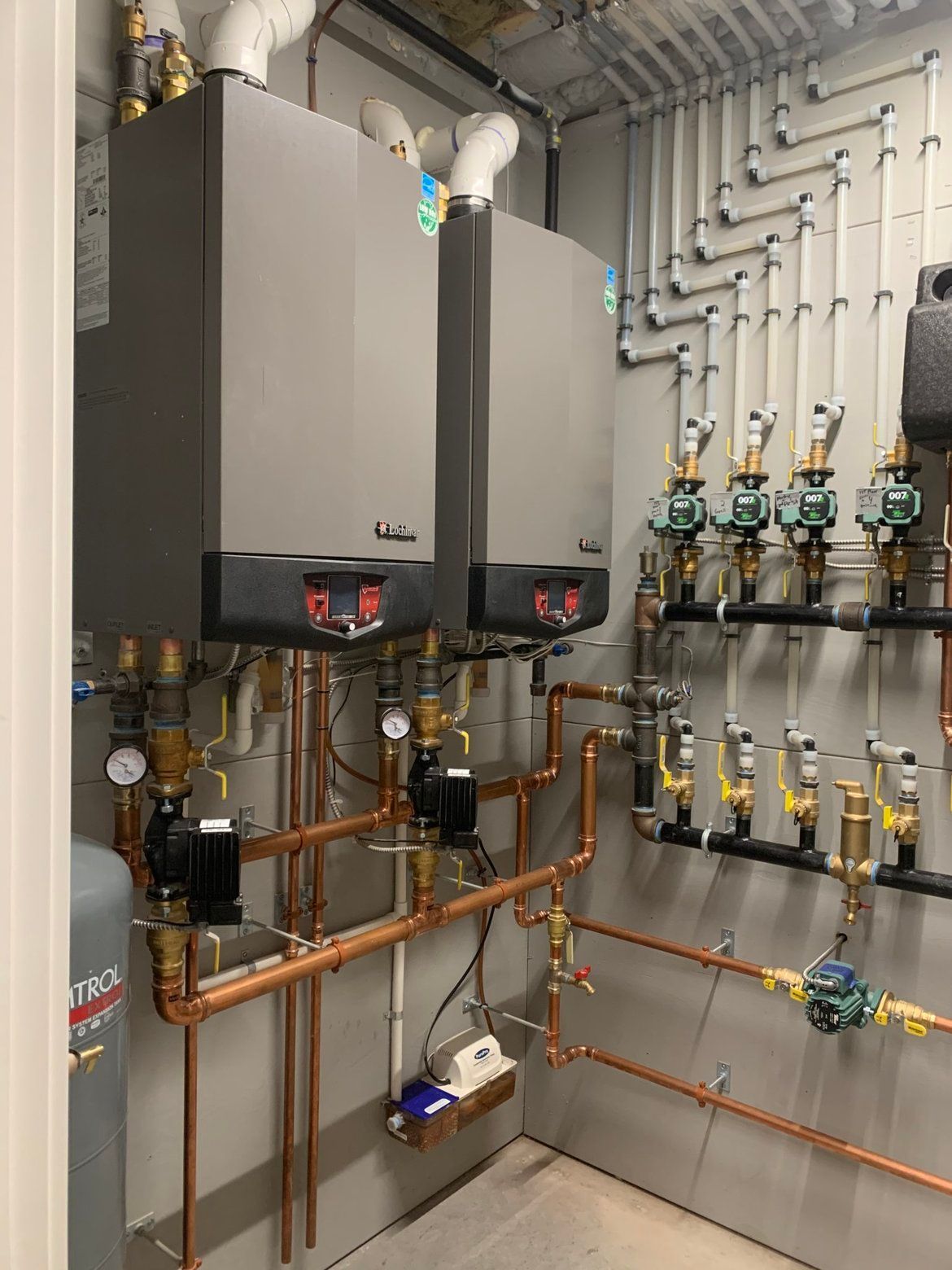Two wall-mounted, gray boilers with copper pipes and valves in an equipment room.