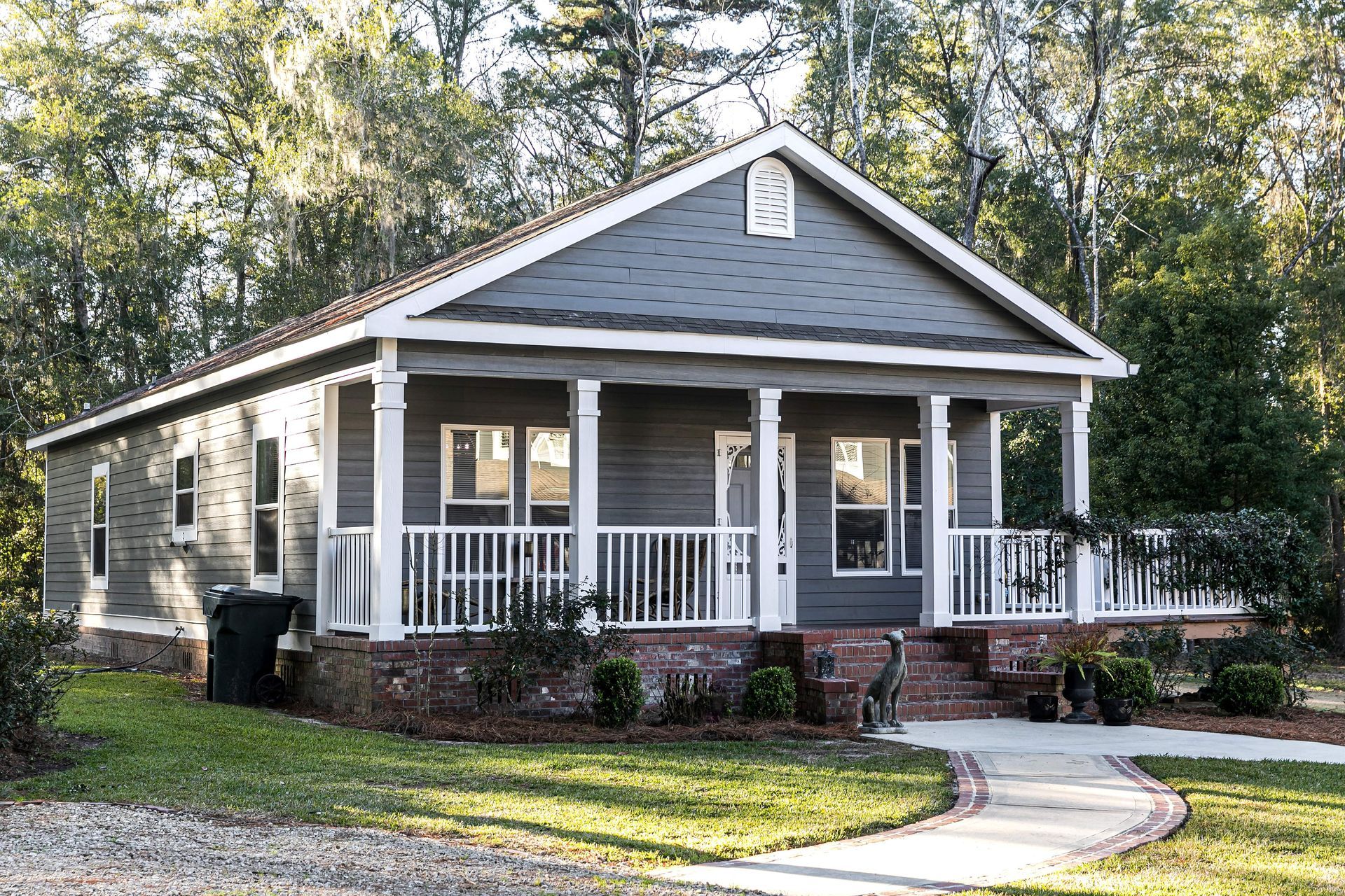A manufactured home with a wide front porch sits on a landscaped lot surrounded by trees.