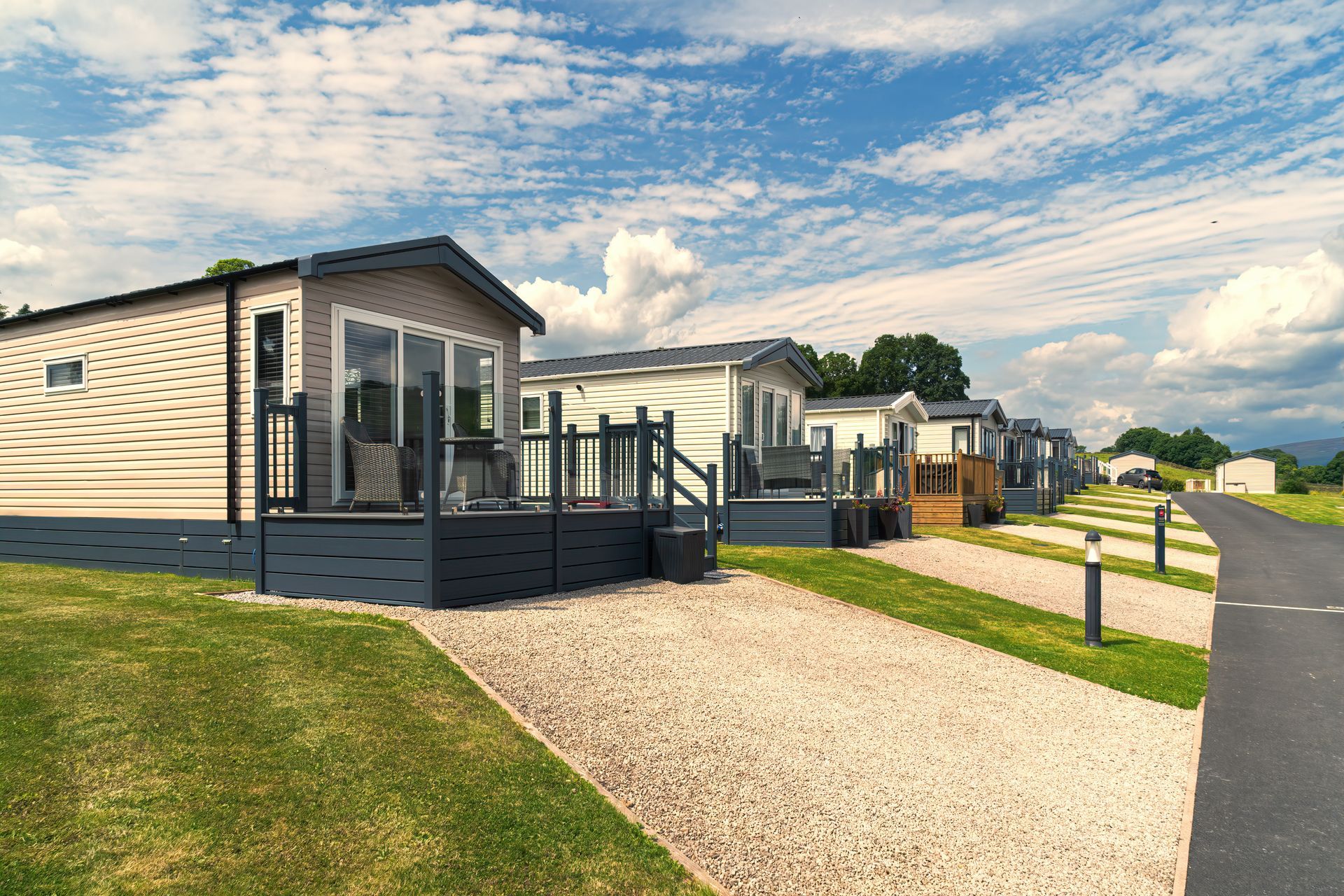 Row of modern holiday caravans with decks along a landscaped park road under a blue sky.