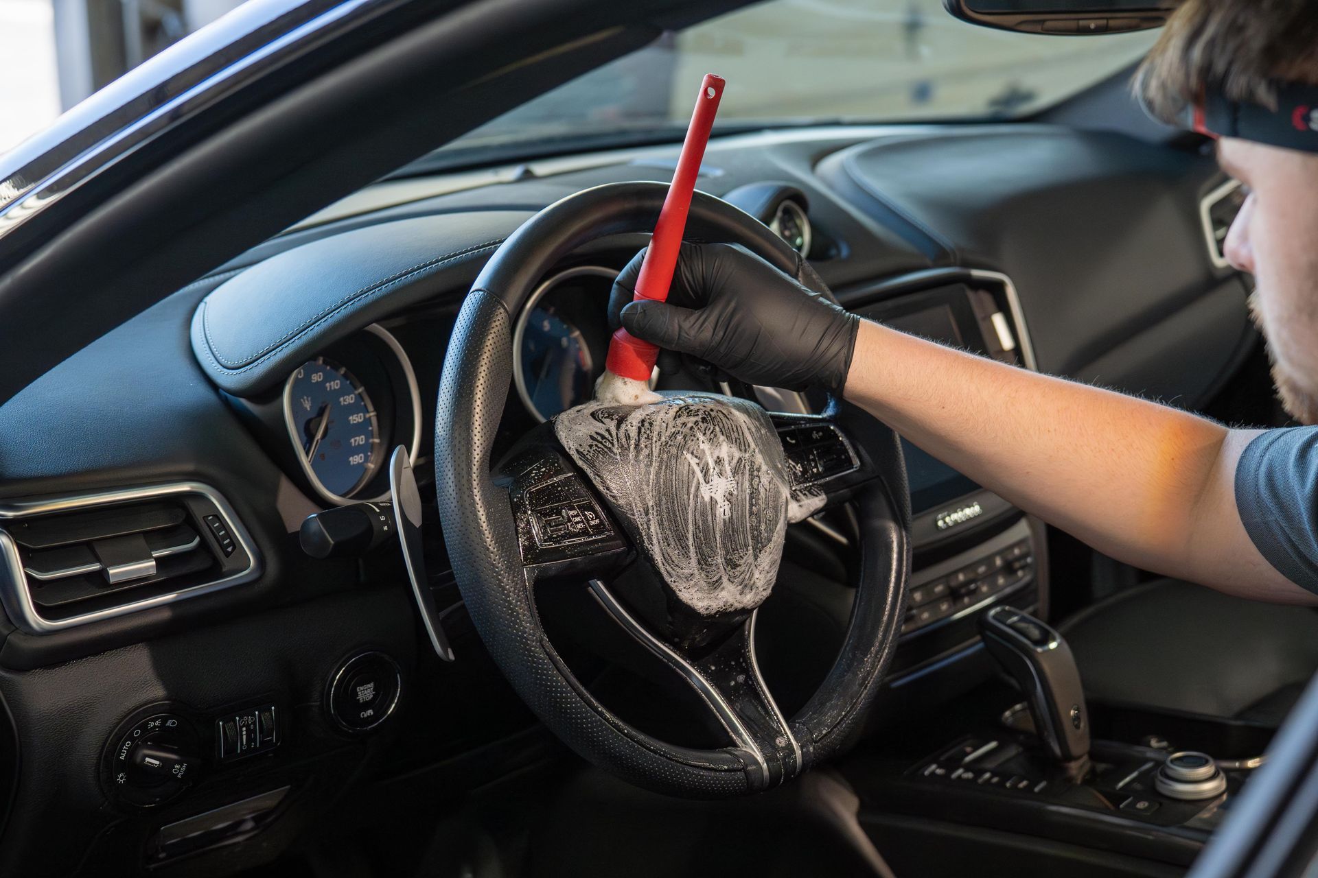 Person cleaning a car's steering wheel with a brush. Interior shot of a car with a black interior and dashboard.