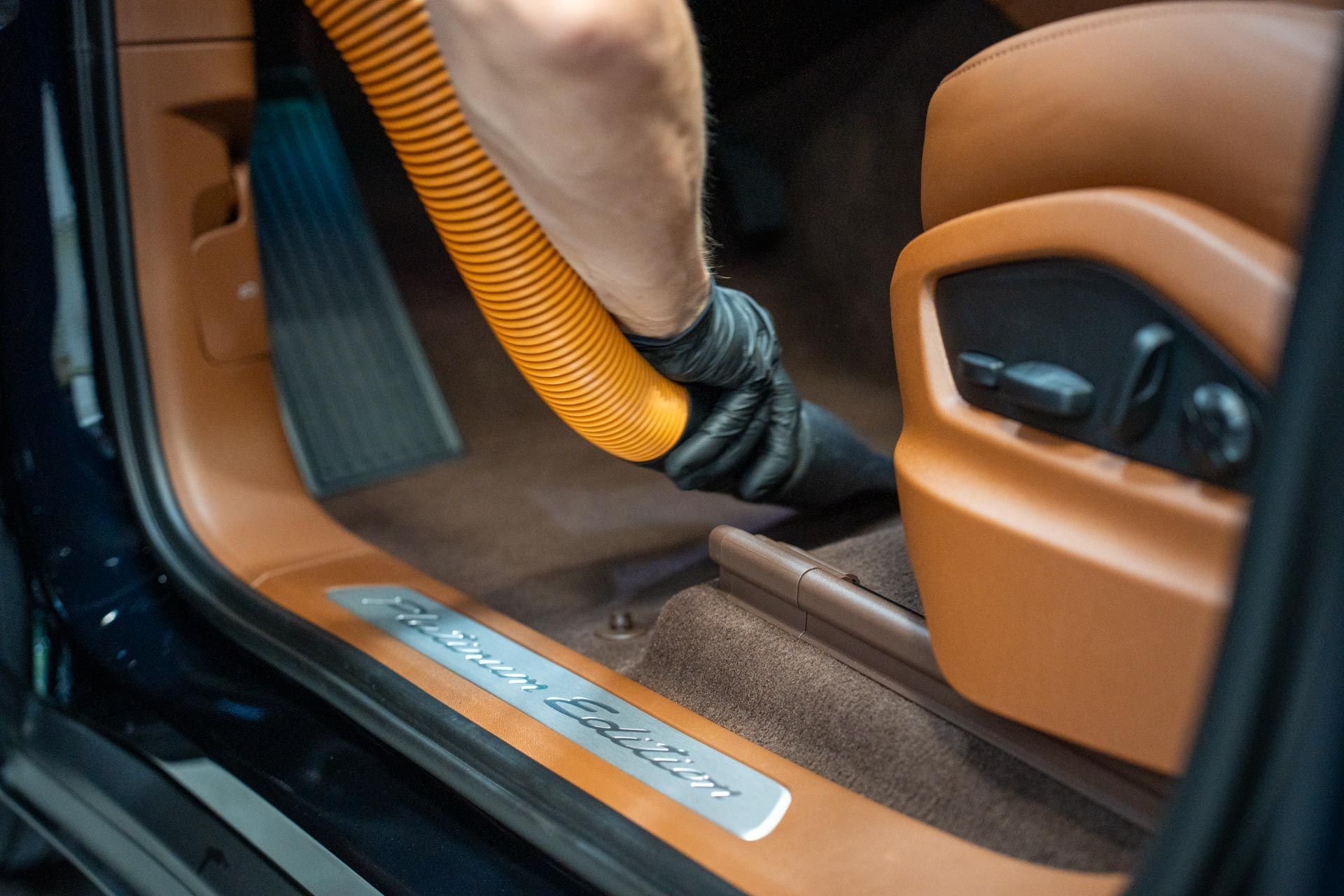 Person vacuuming the car floor, with orange hose and brown leather interior.