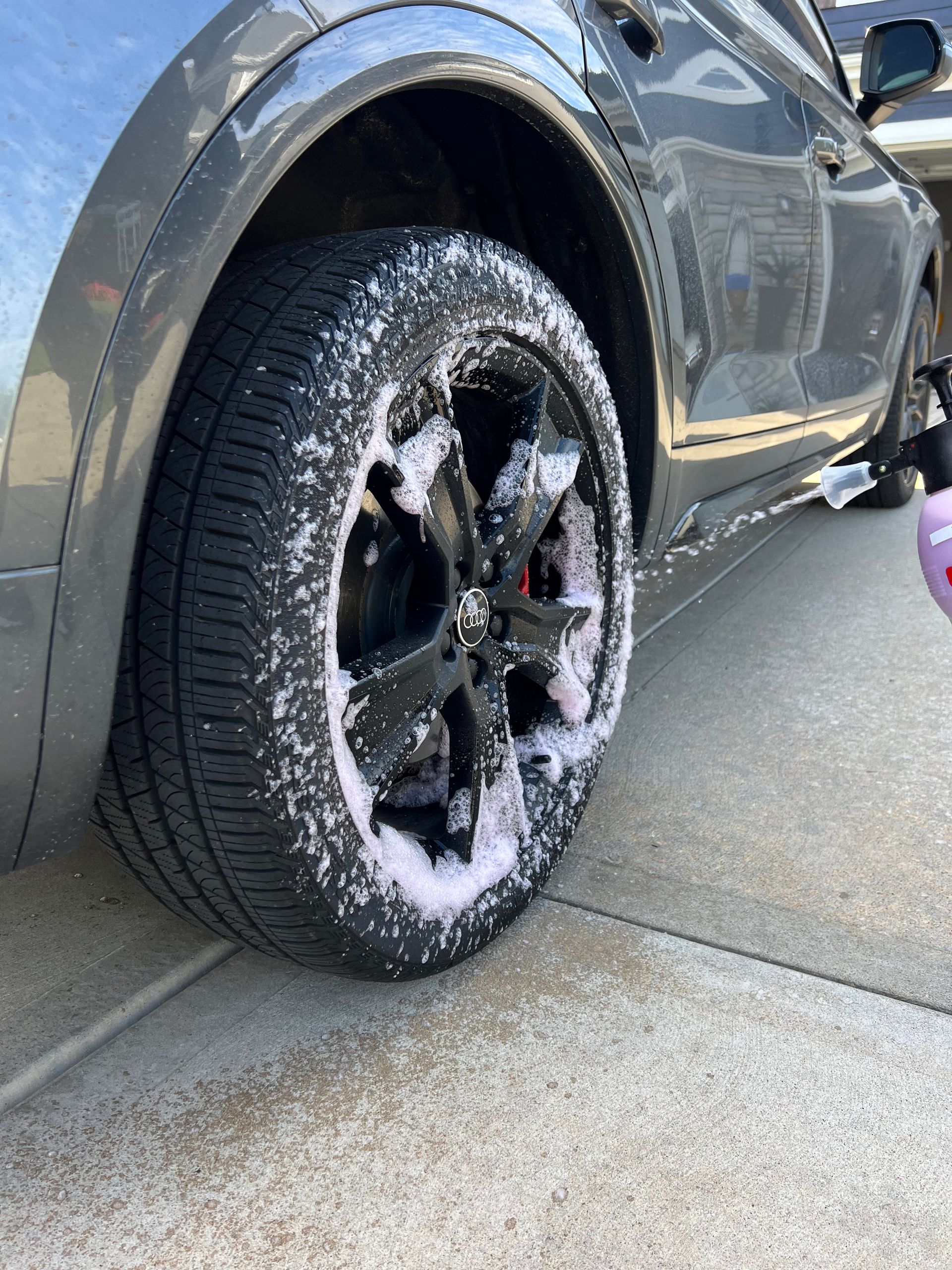 A gray SUV wheel being sprayed with pink car wash foam. Black wheel rim and tire visible.