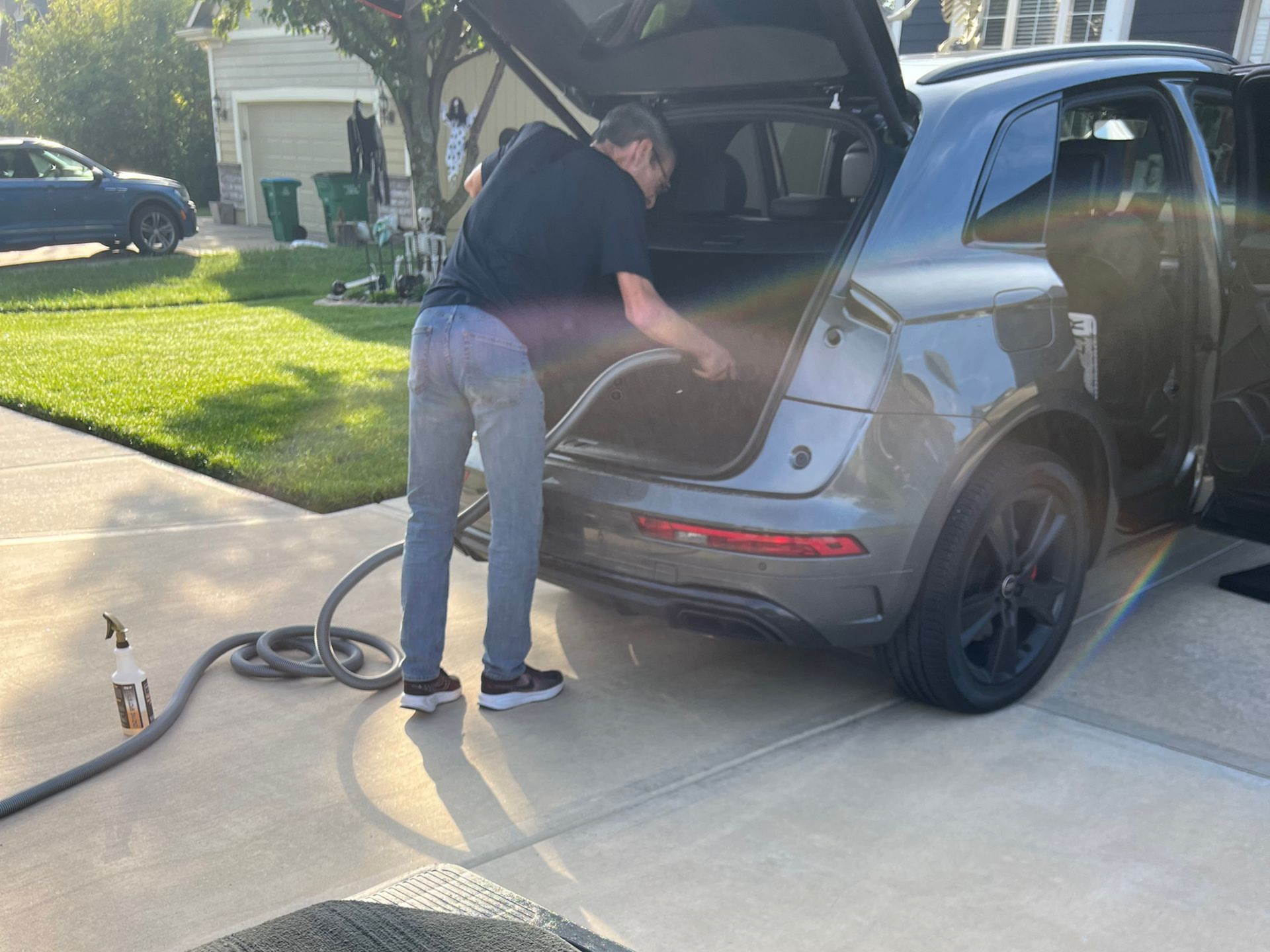 Man vacuuming the trunk of a gray SUV on a driveway.