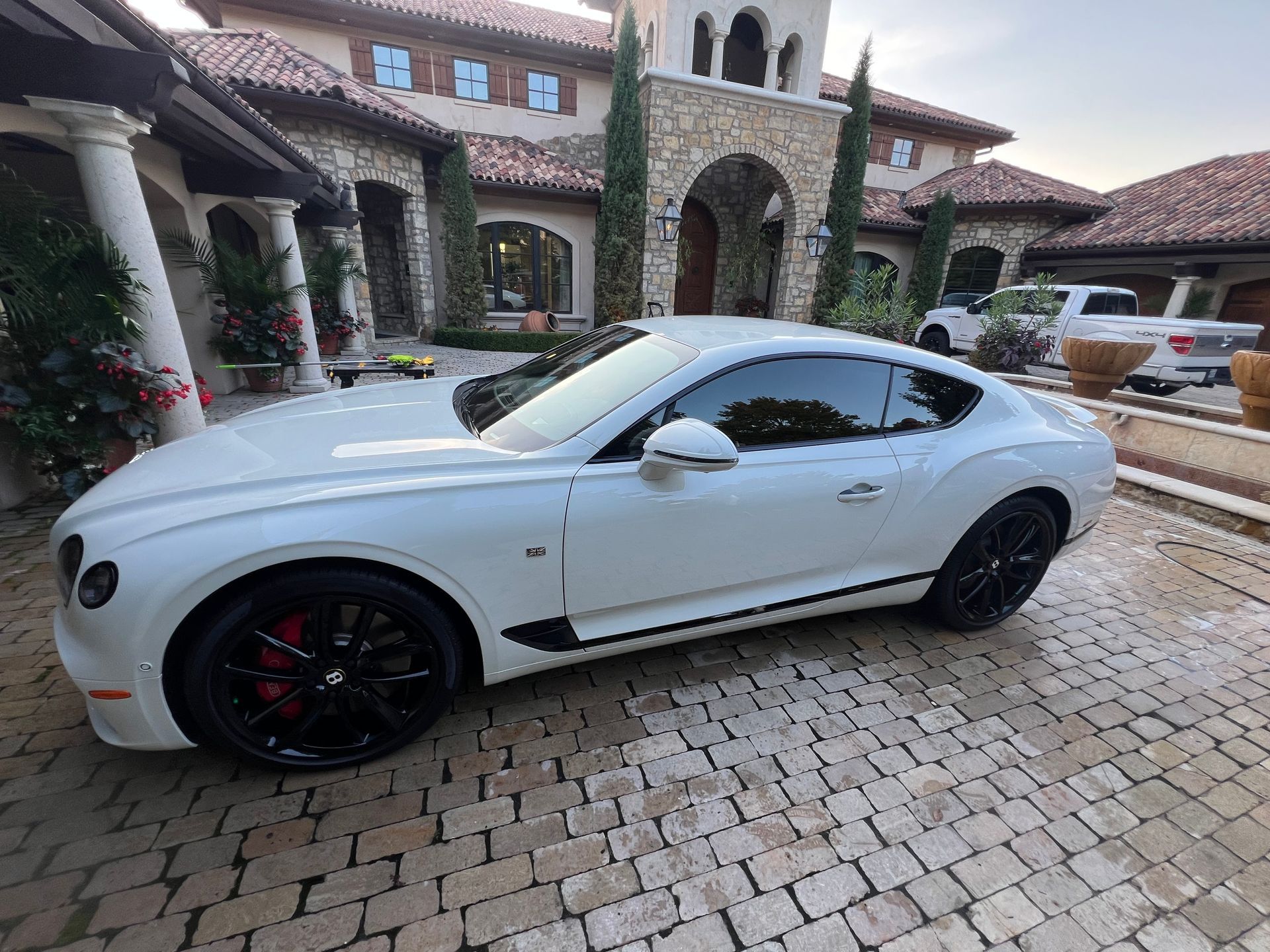 White Bentley coupe parked on a brick driveway in front of a large house.