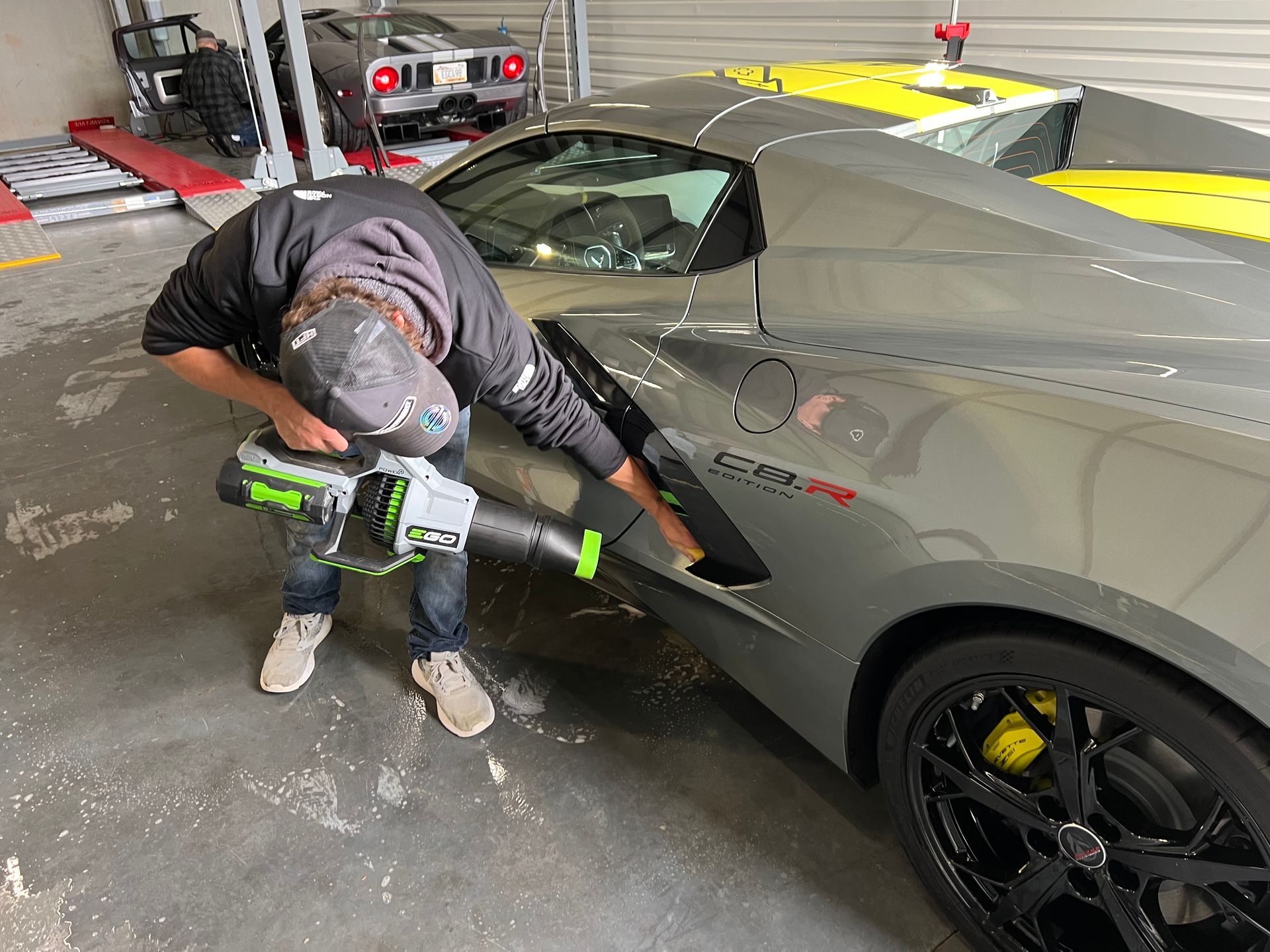 A person uses a leaf blower to clean a gray sports car in a garage.