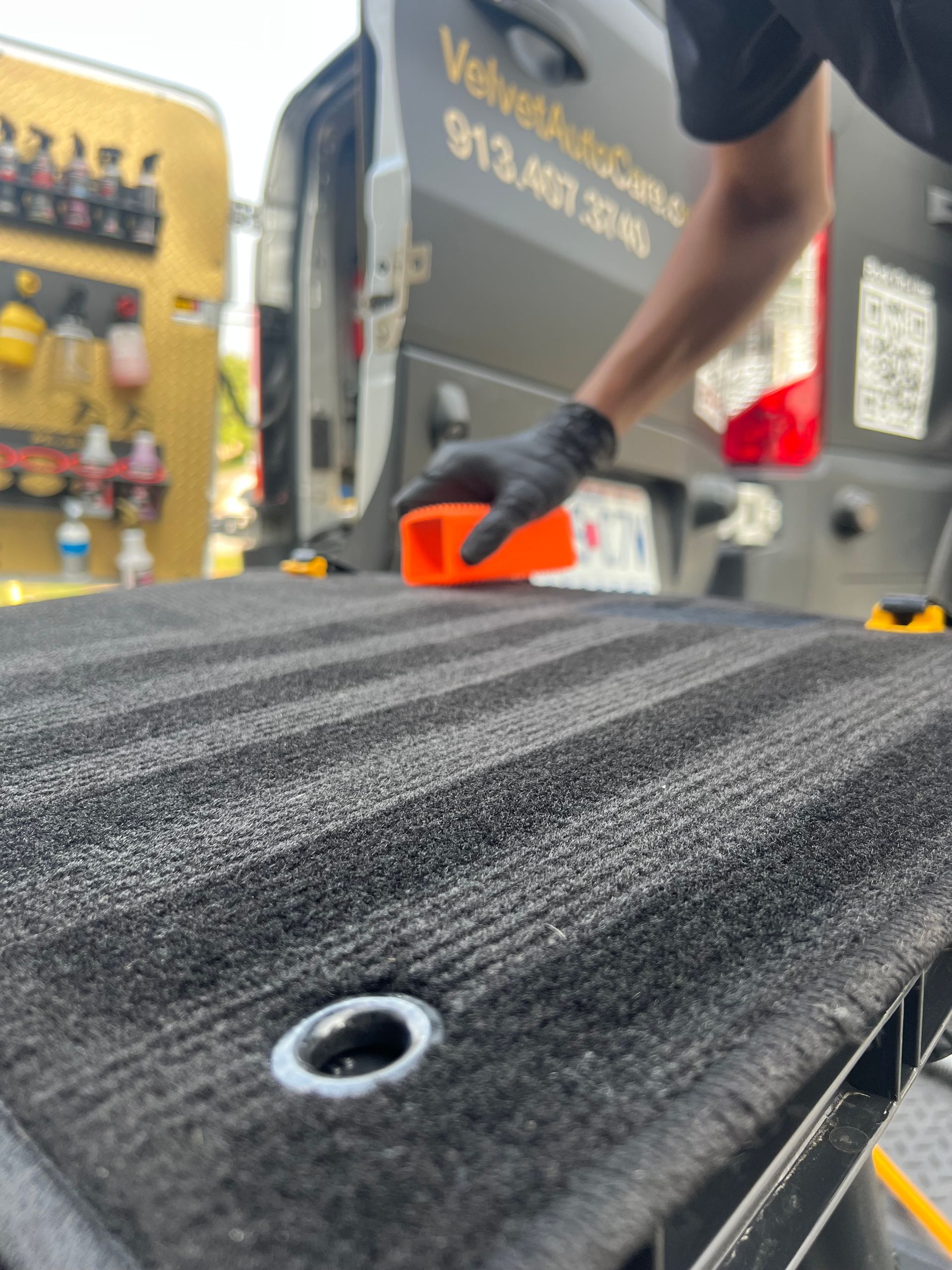 A person wearing black gloves cleans a black truck bed. An orange block is used for scrubbing.