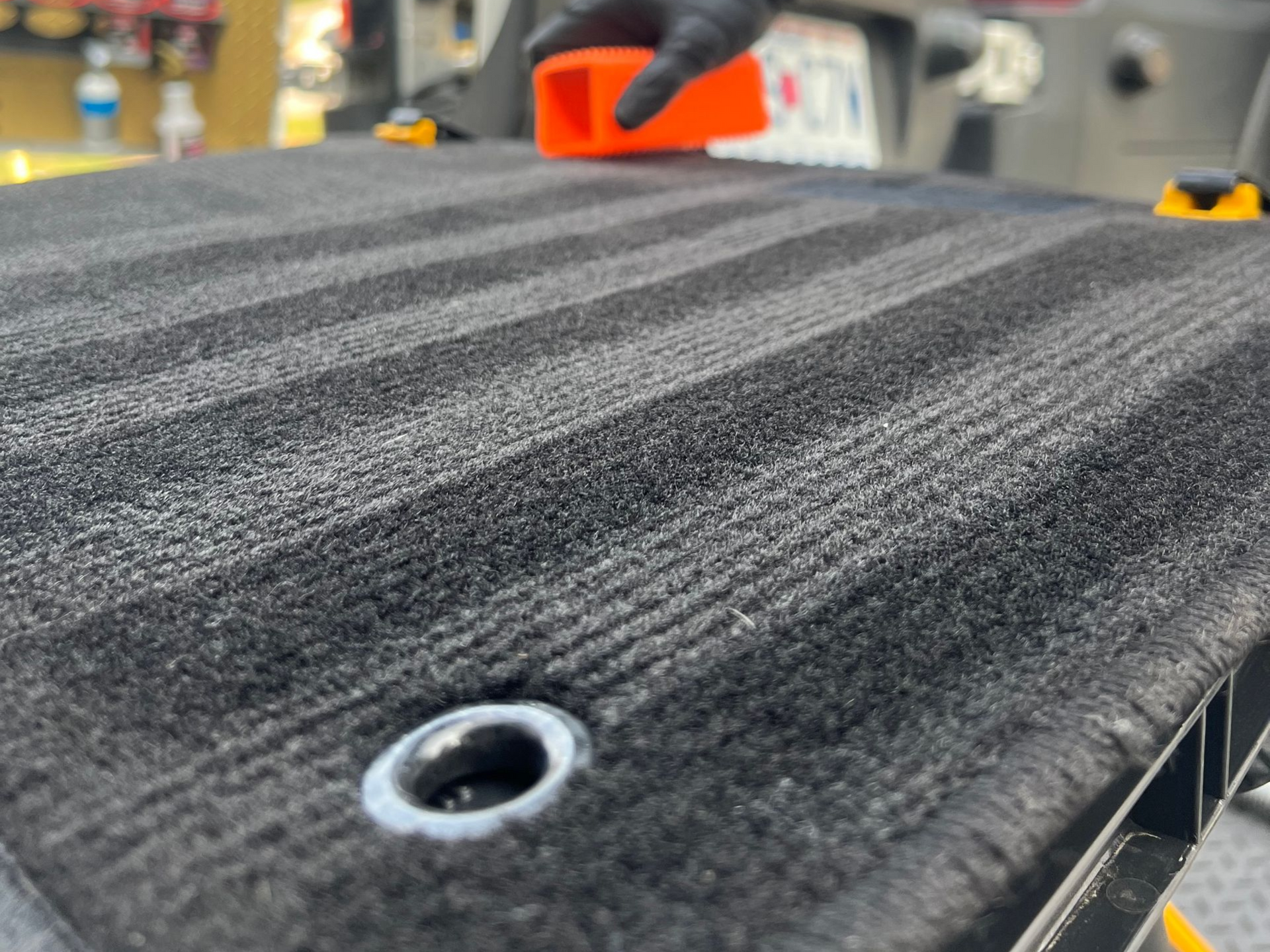 Person in black glove using orange tool to clean black car mat with horizontal grooves.
