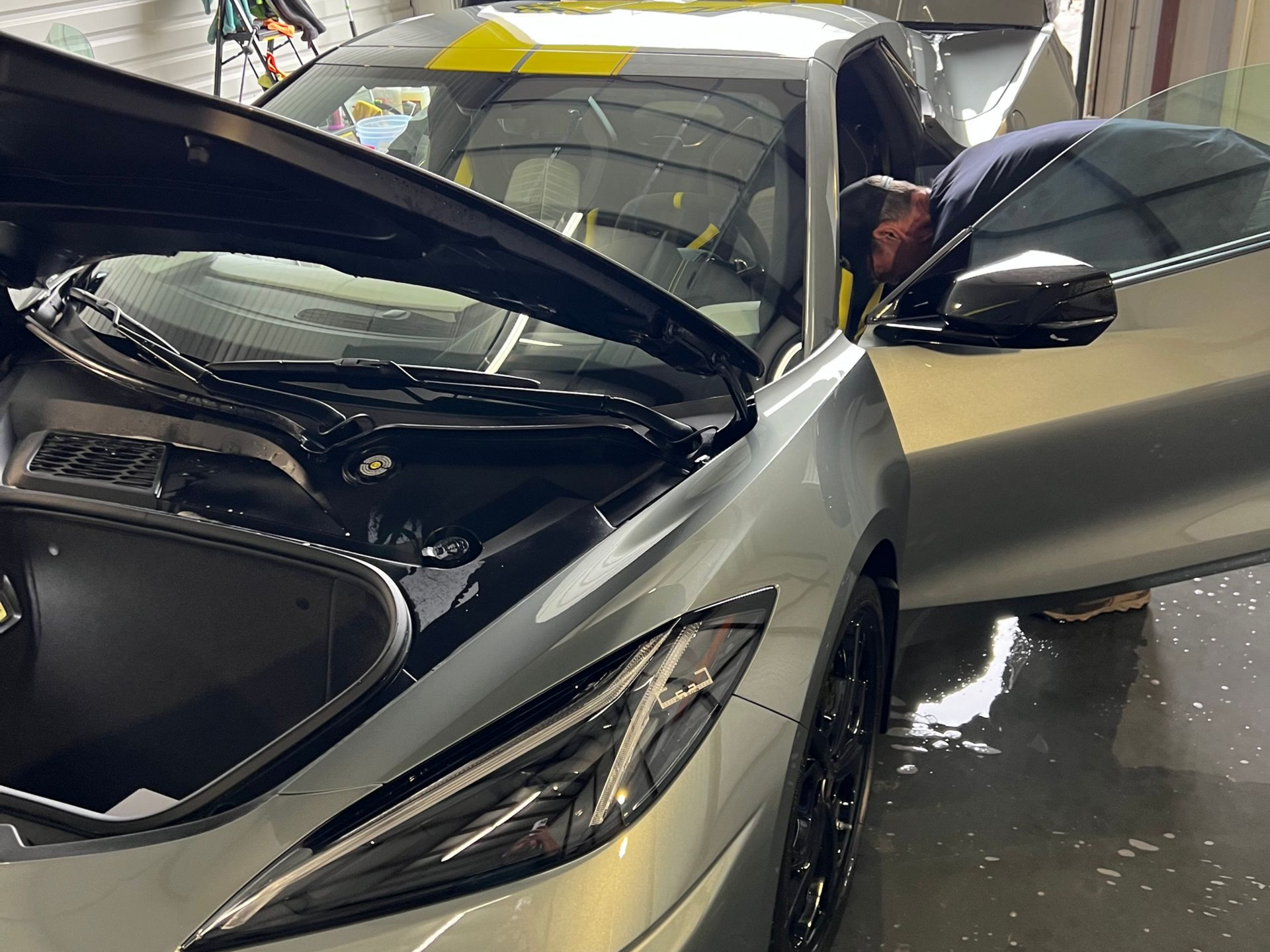 A person cleaning a silver sports car with the hood and door open. Yellow stripe on top.