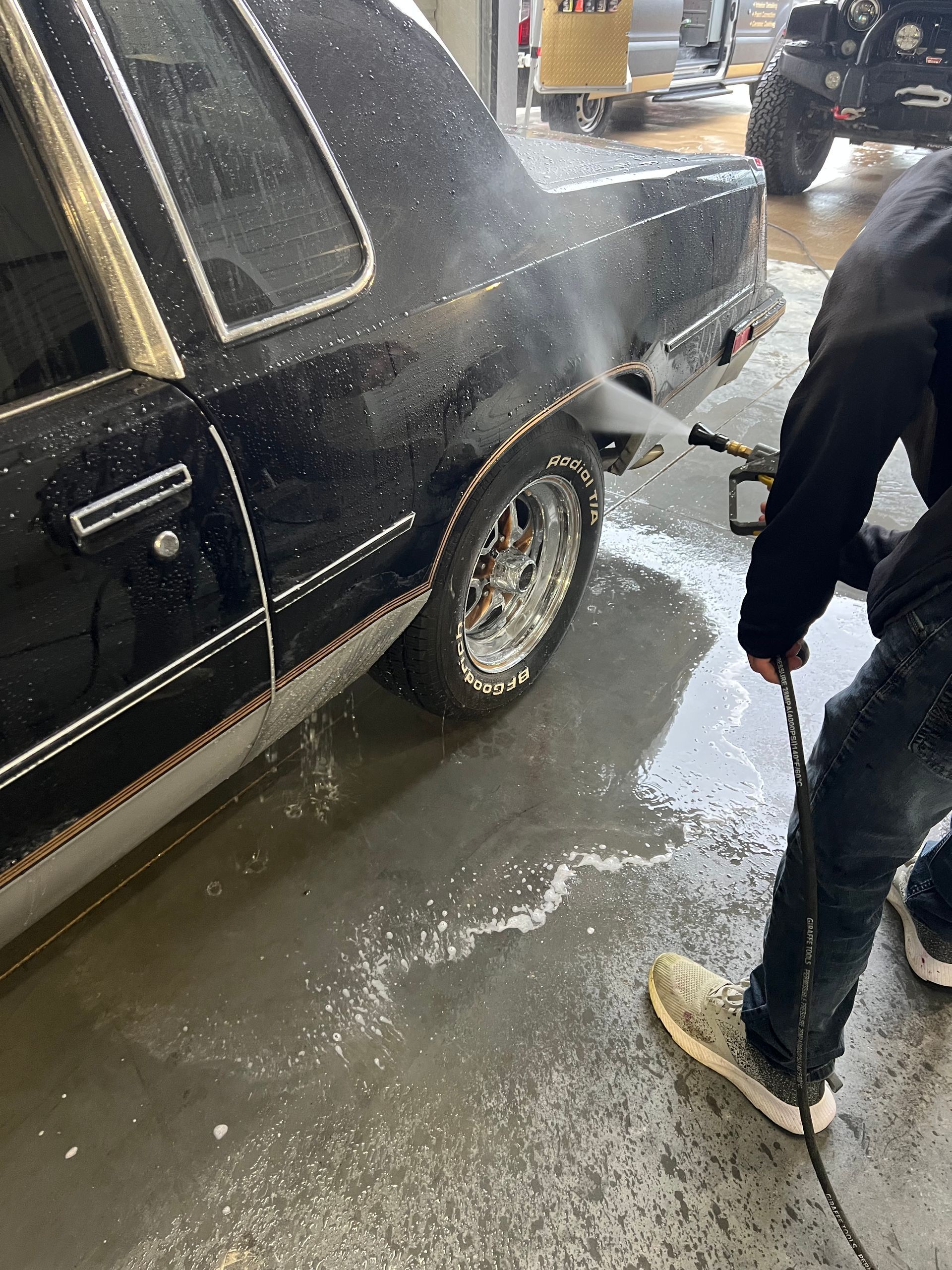 Person power washing a black car with chrome wheels. Concrete floor.