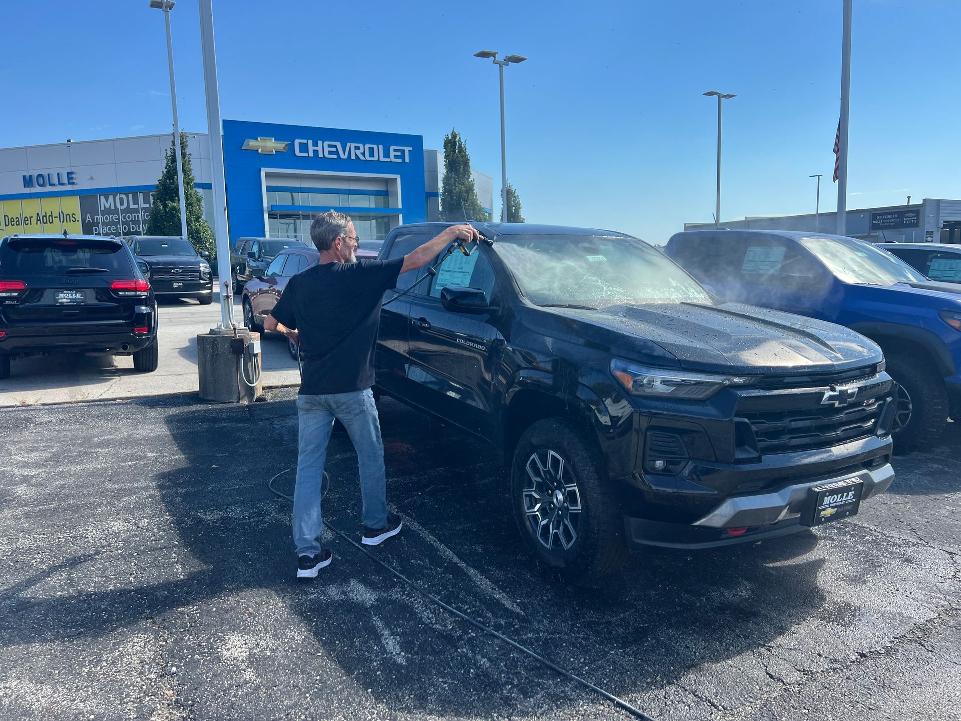 Man washing a black Chevrolet truck at a car dealership on a sunny day.