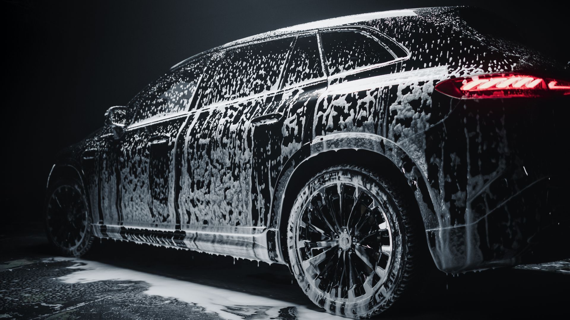 Black SUV covered in white foam, illuminated with red taillights, washing outdoors at night.