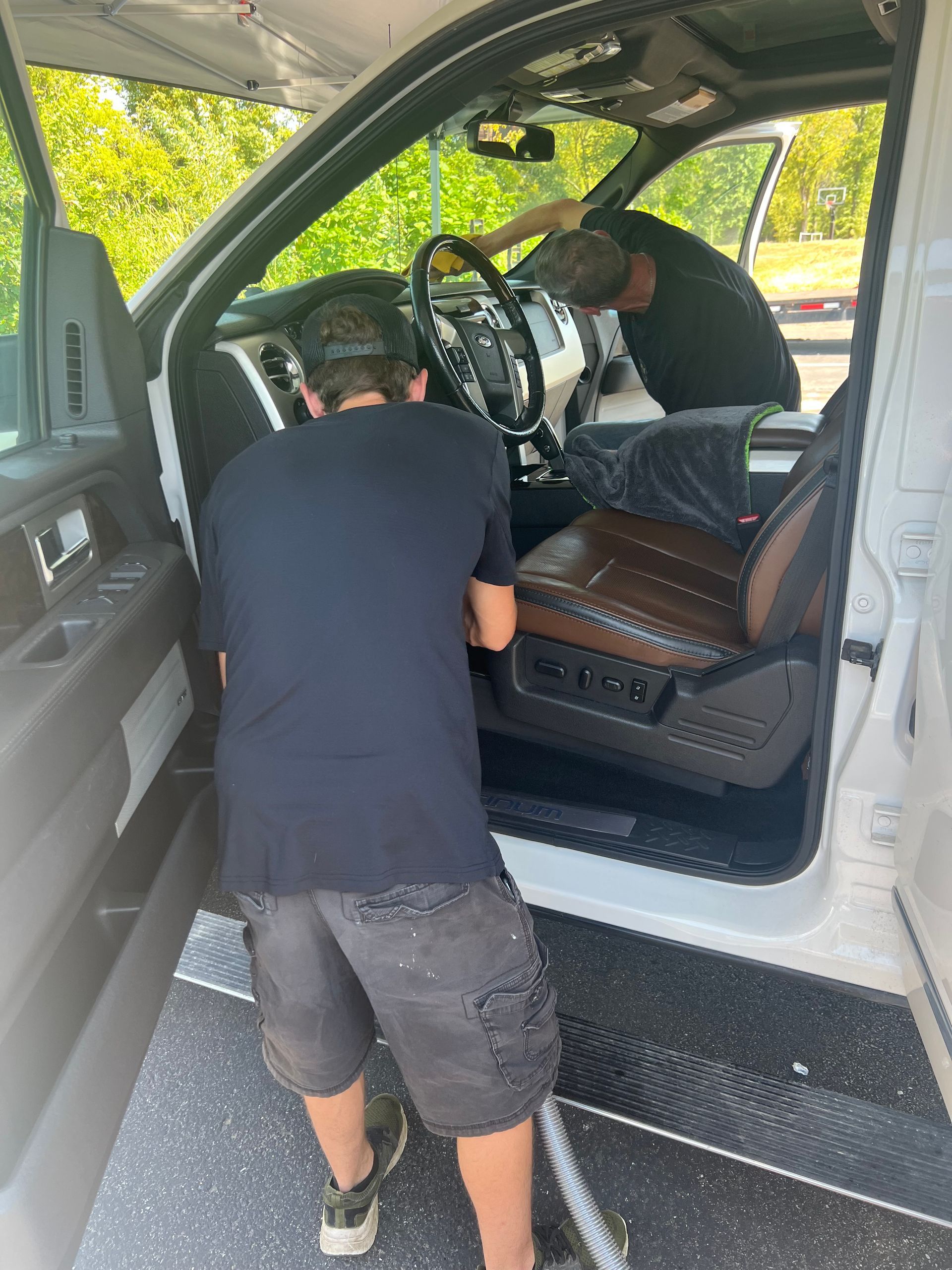 Two people cleaning the interior of a white truck. One person in a black shirt, another visible working near the steering wheel.