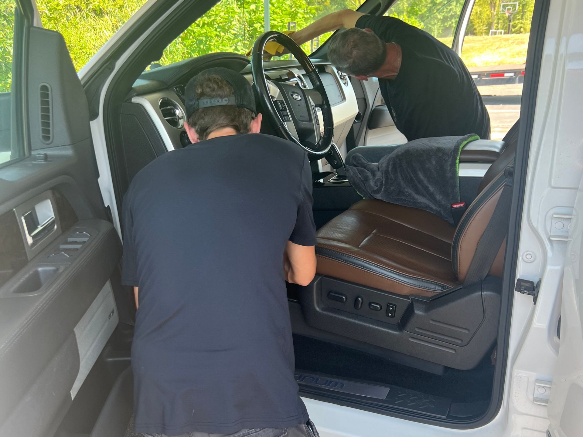 Two people cleaning the interior of a white truck. One cleans the steering wheel, the other the dashboard.