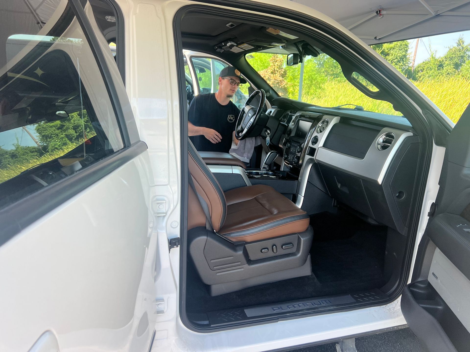 Man sitting inside a white truck, door open. Brown and black interior. Bright outside.