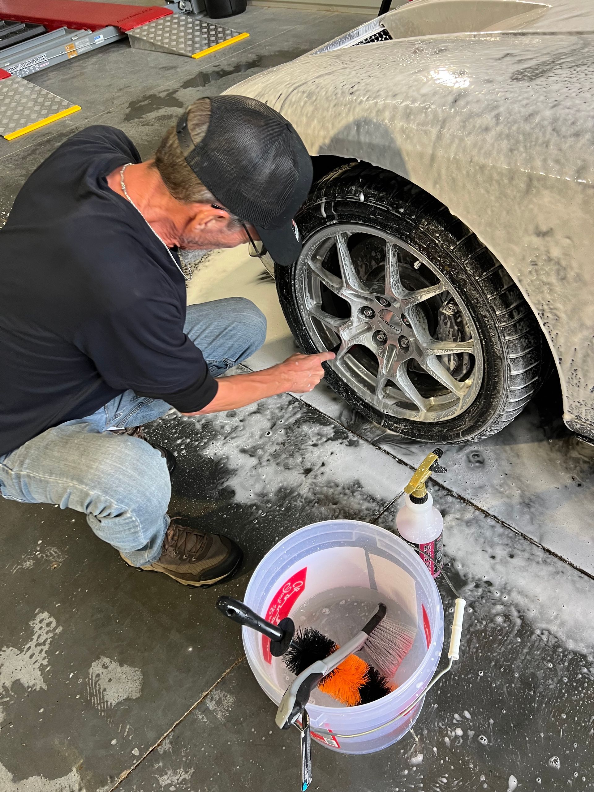Man washing a car tire with a brush and soapy water. A bucket of supplies sits nearby.