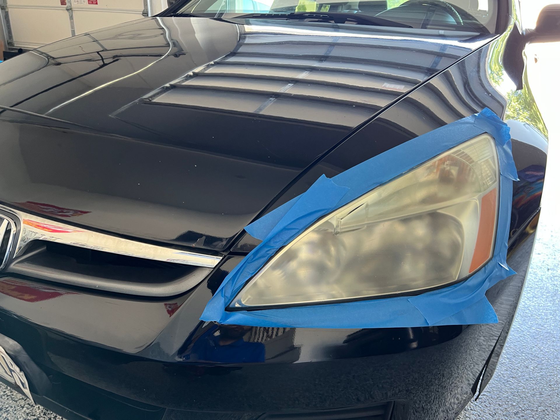 A black car with blue tape around a cloudy headlight, garage setting.