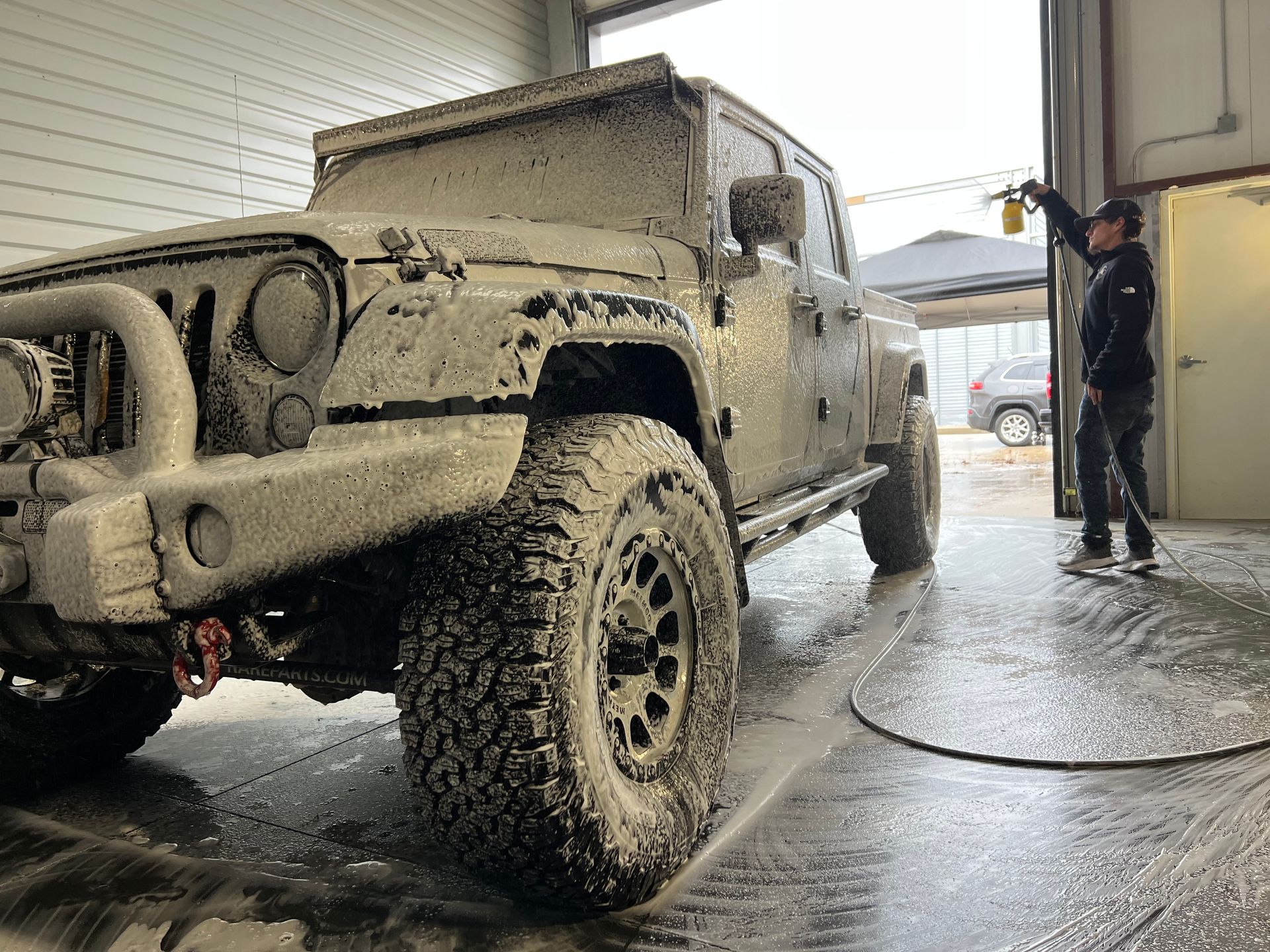 Jeep covered in foam at a car wash. A person sprays the vehicle with a hose.