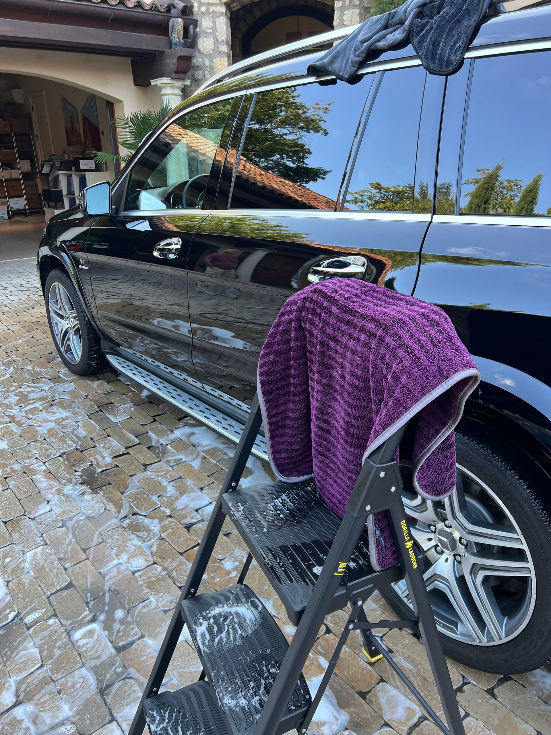 Black SUV being washed; purple towel on a step ladder. Bubbles on the paved driveway.