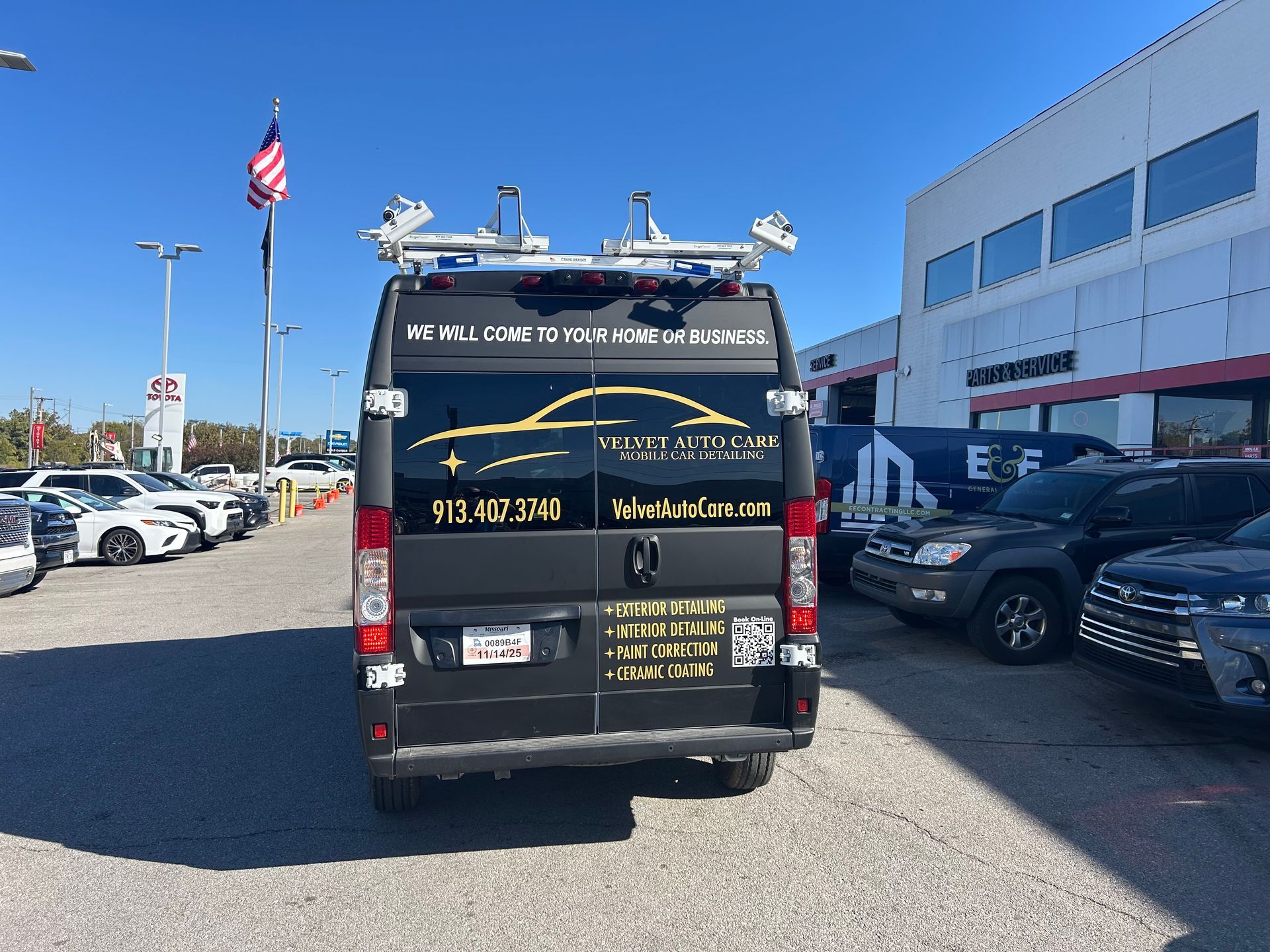 Black van with ladder rack parked at a dealership. Text on the back includes a phone number and business name.
