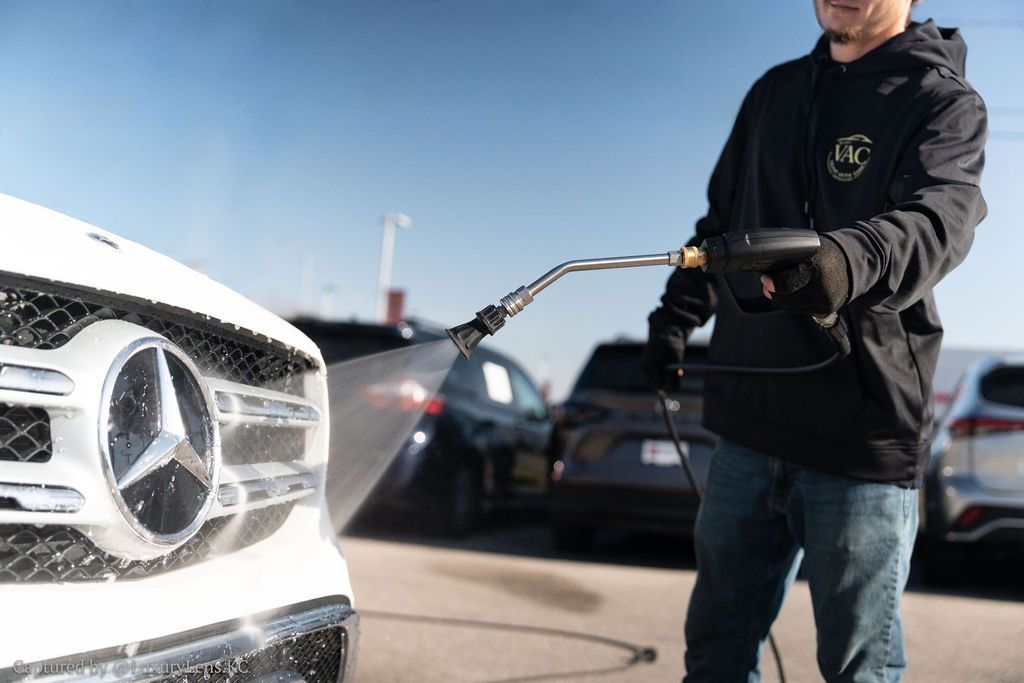 Man washing a white Mercedes-Benz with a pressure washer outdoors; soapy spray, sunny day.