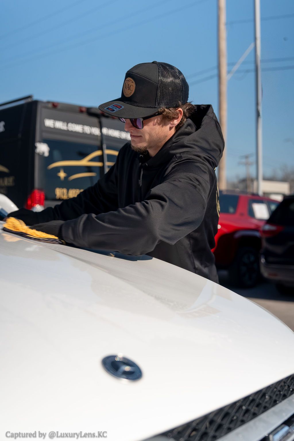 Person in black hoodie and hat wiping a white car with a yellow cloth outdoors.
