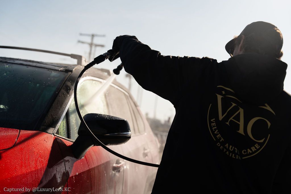 Person washing a red car with a pressure washer outdoors; bright sunny day.