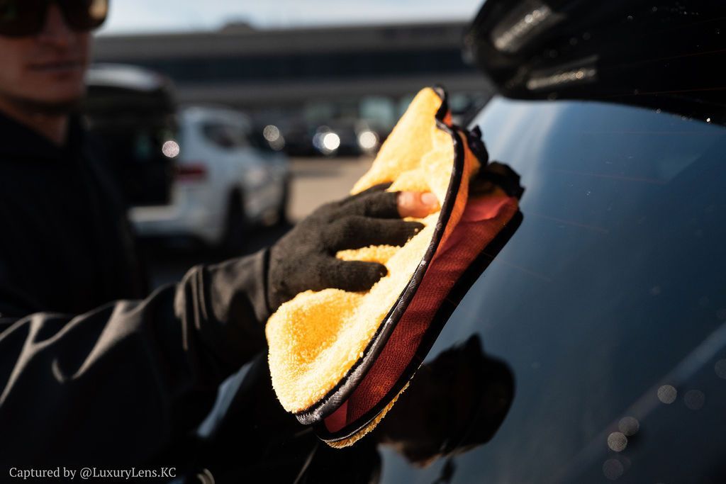 Person in black gloves wiping a car's black surface with a yellow and red microfiber cloth.