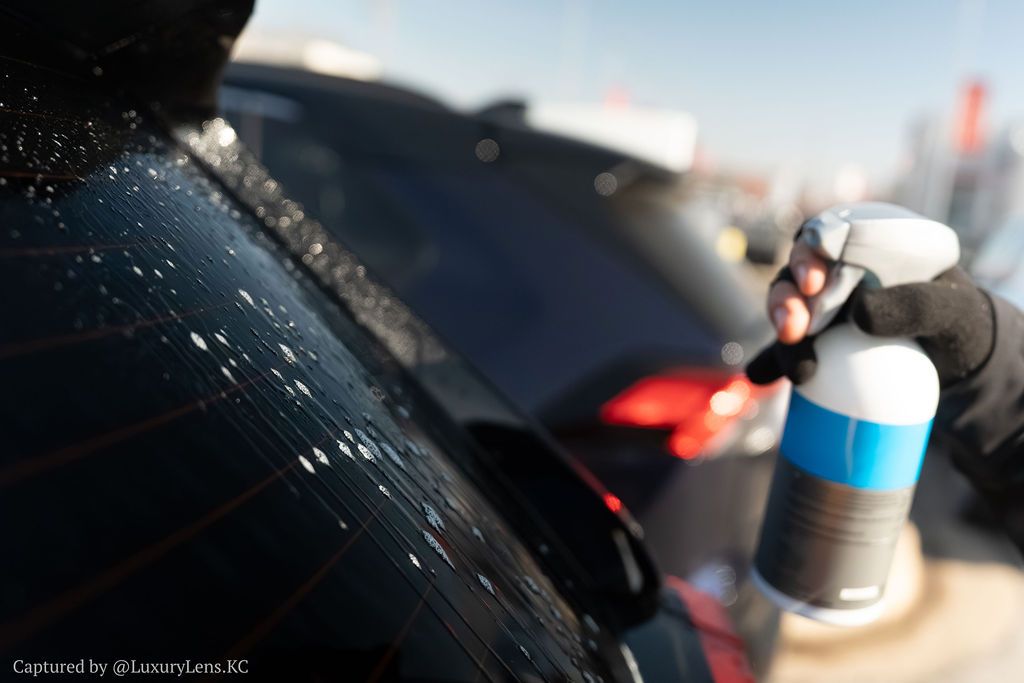 Person sprays water or cleaner on the rear window of a black car with a blue and white spray bottle.