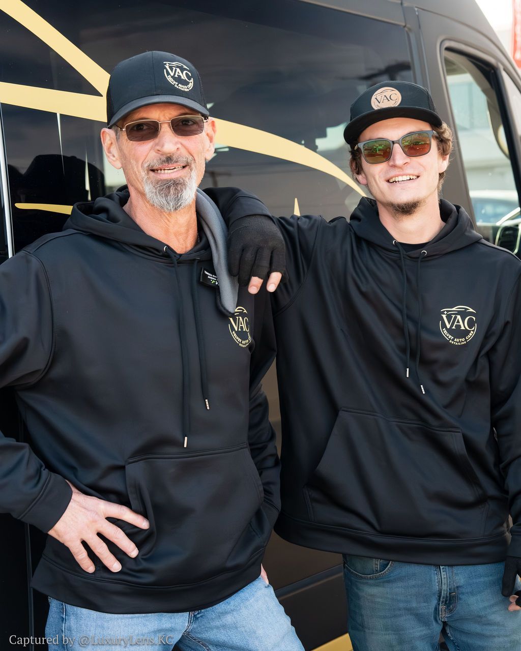Two men in black hoodies and caps pose in front of a van. One has a hand on his hip, the other rests his arm on the first man's shoulder.