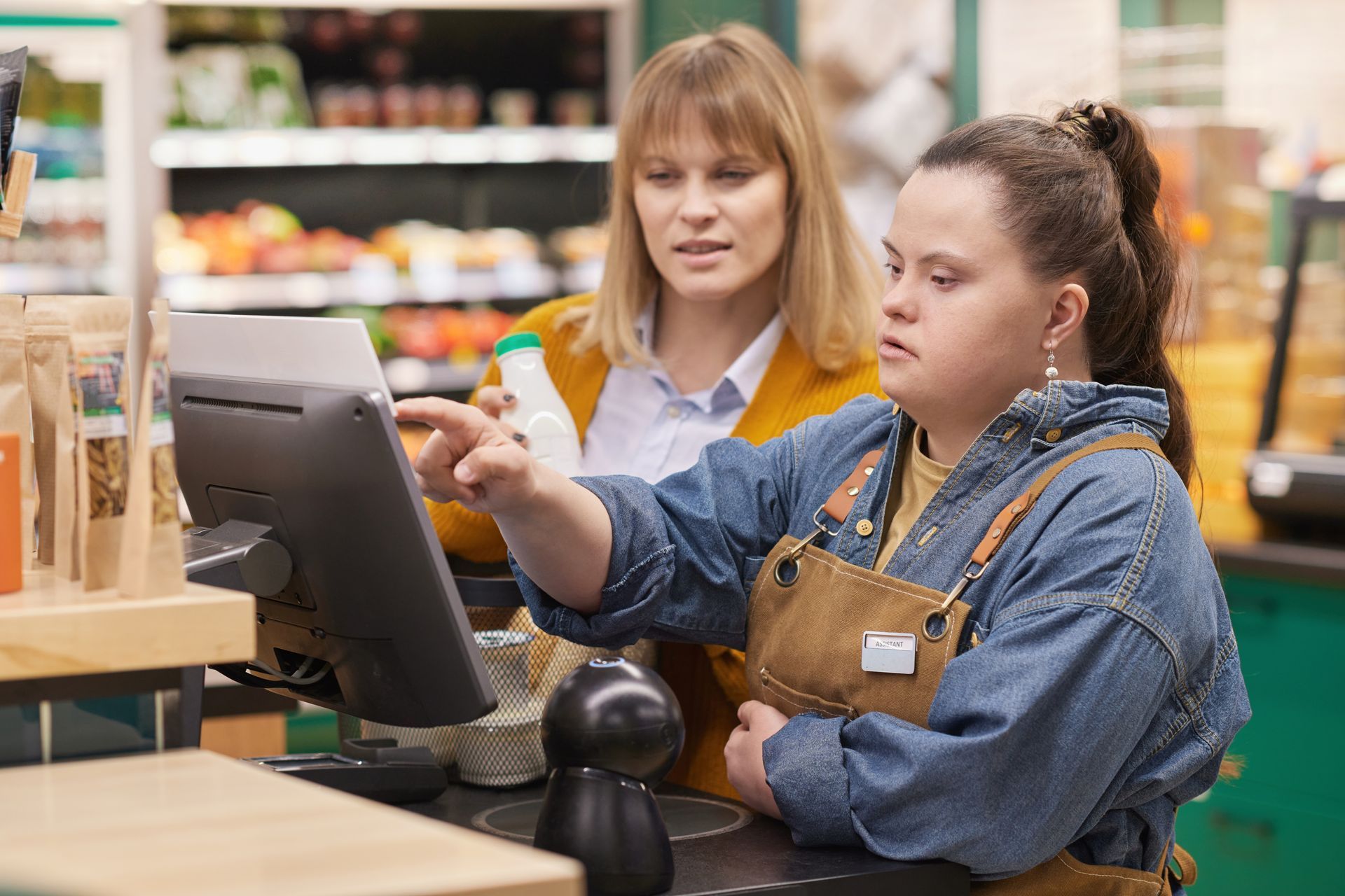 A woman is helping a woman with down syndrome use a computer in a grocery store.