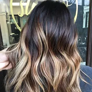 A woman with long dark hair with blonde highlights is standing in front of a window.