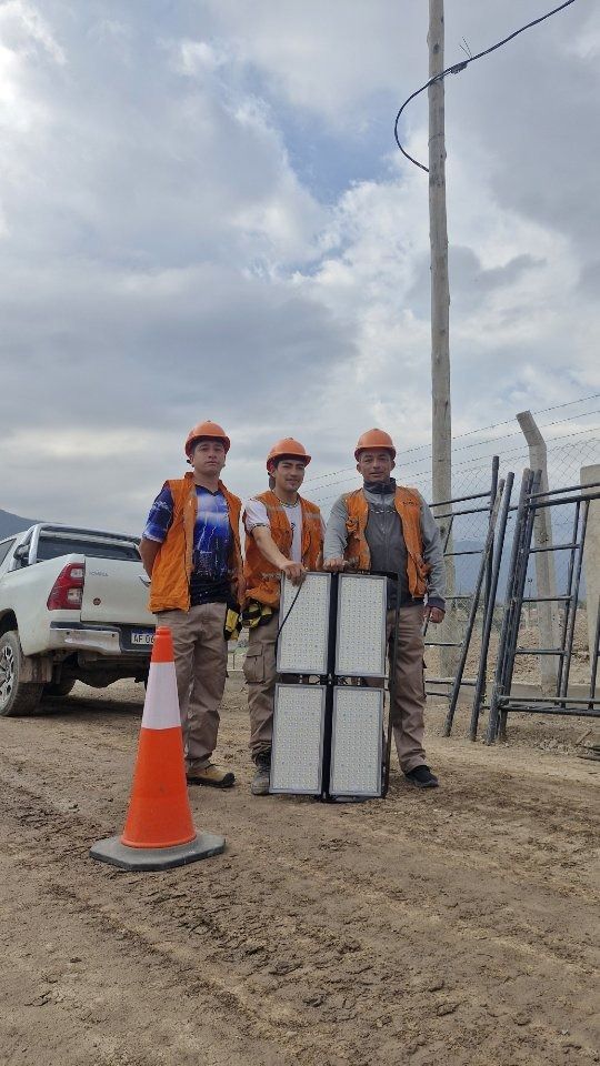 Tres trabajadores con chalecos naranjas y cascos sostienen un objeto grande y rectangular en un camino de tierra.