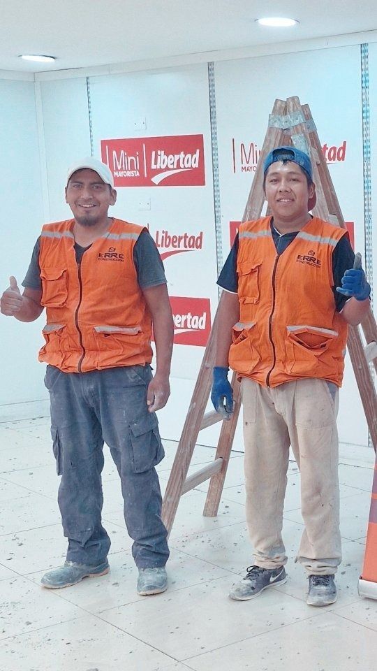 Dos trabajadores con chalecos naranjas posan cerca de una escalera y un cartel de 