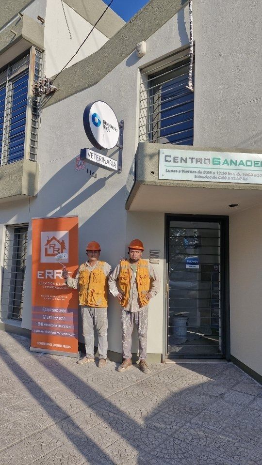 Dos trabajadores con chalecos antibalas se encuentran junto a la entrada de un edificio. A la izquierda hay una pancarta.