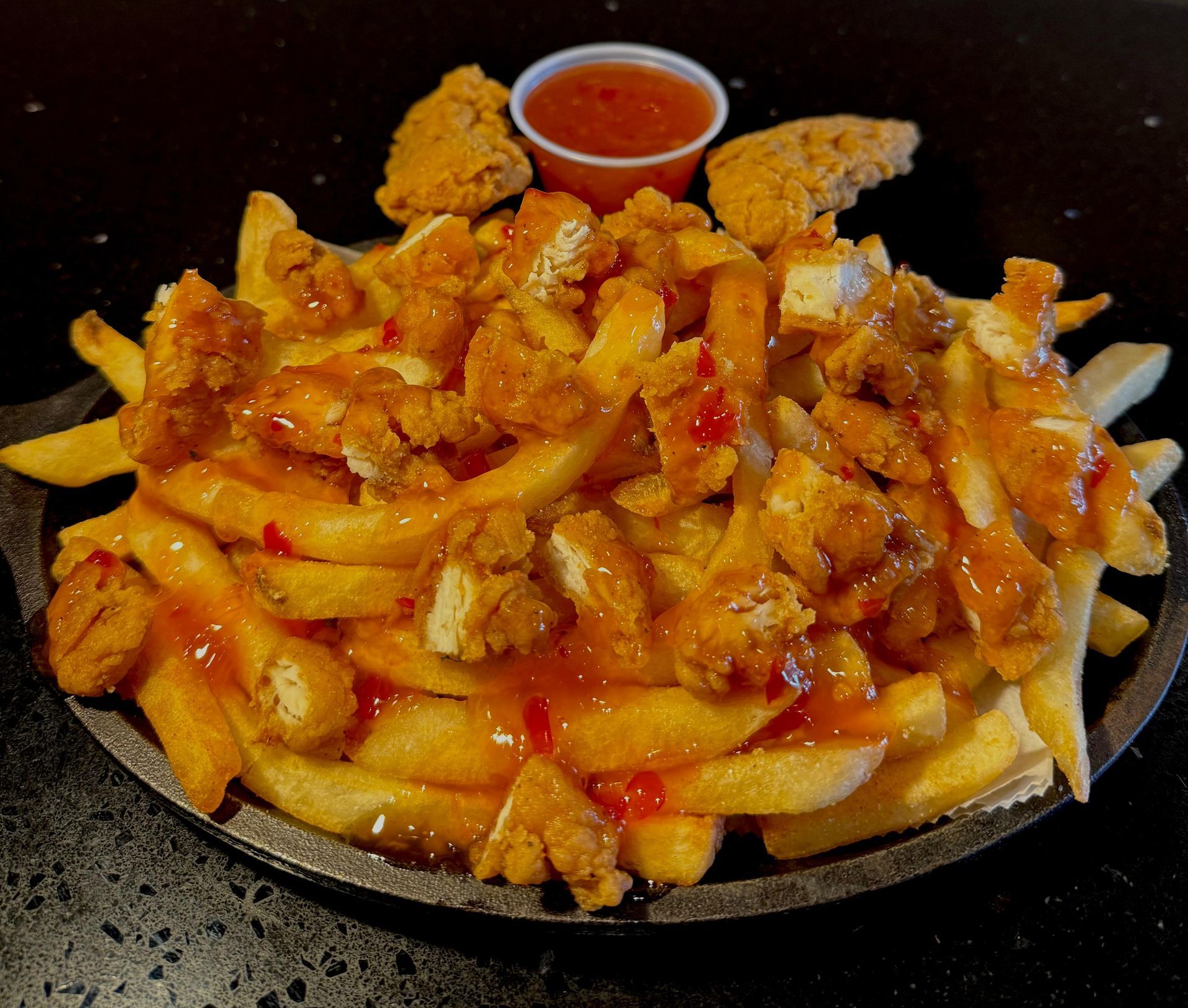 Fries topped with fried chicken pieces and sauce, served with a small side of sauce.