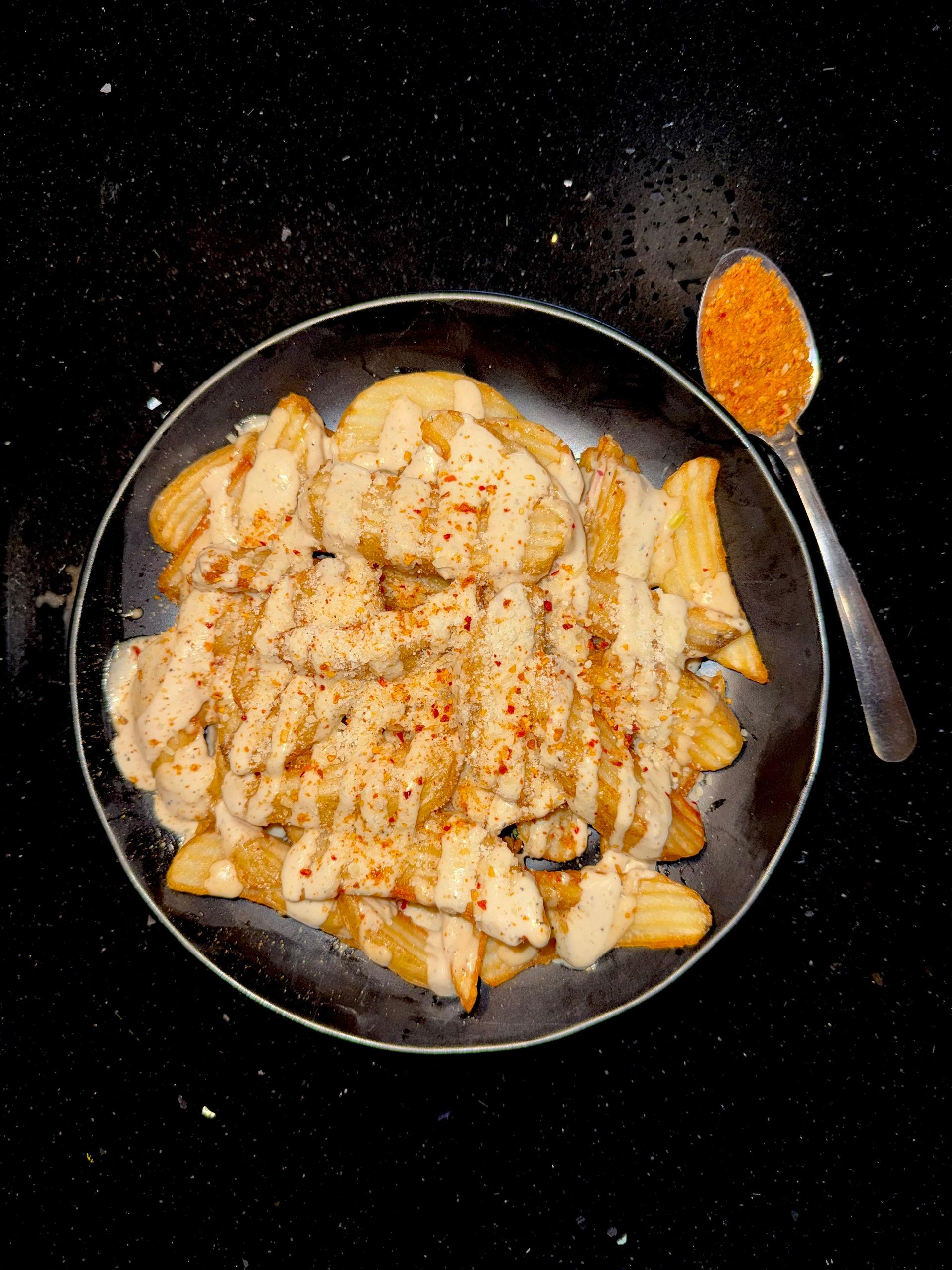 Potato wedges with creamy sauce and spice, served on a black plate with a spoon of spice.