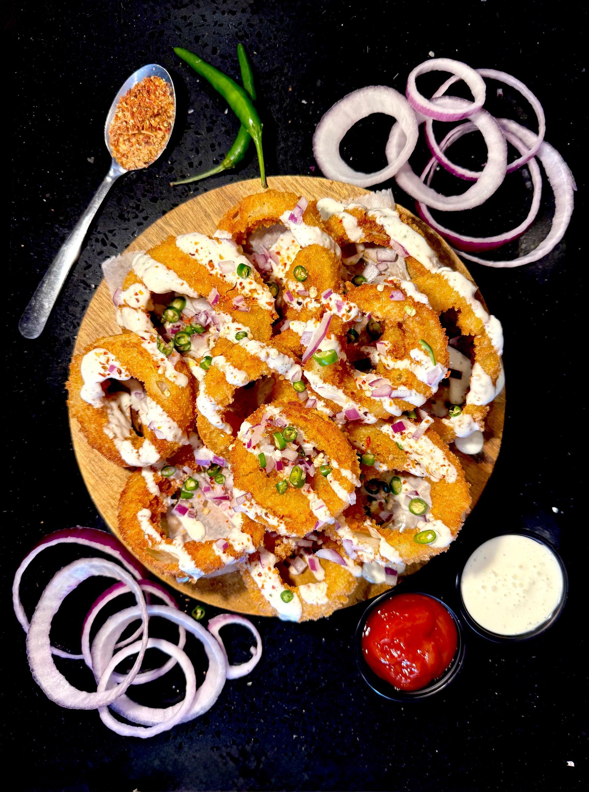 Fried onion rings with sauce and spices on a wooden board, garnished with onions and green chili.
