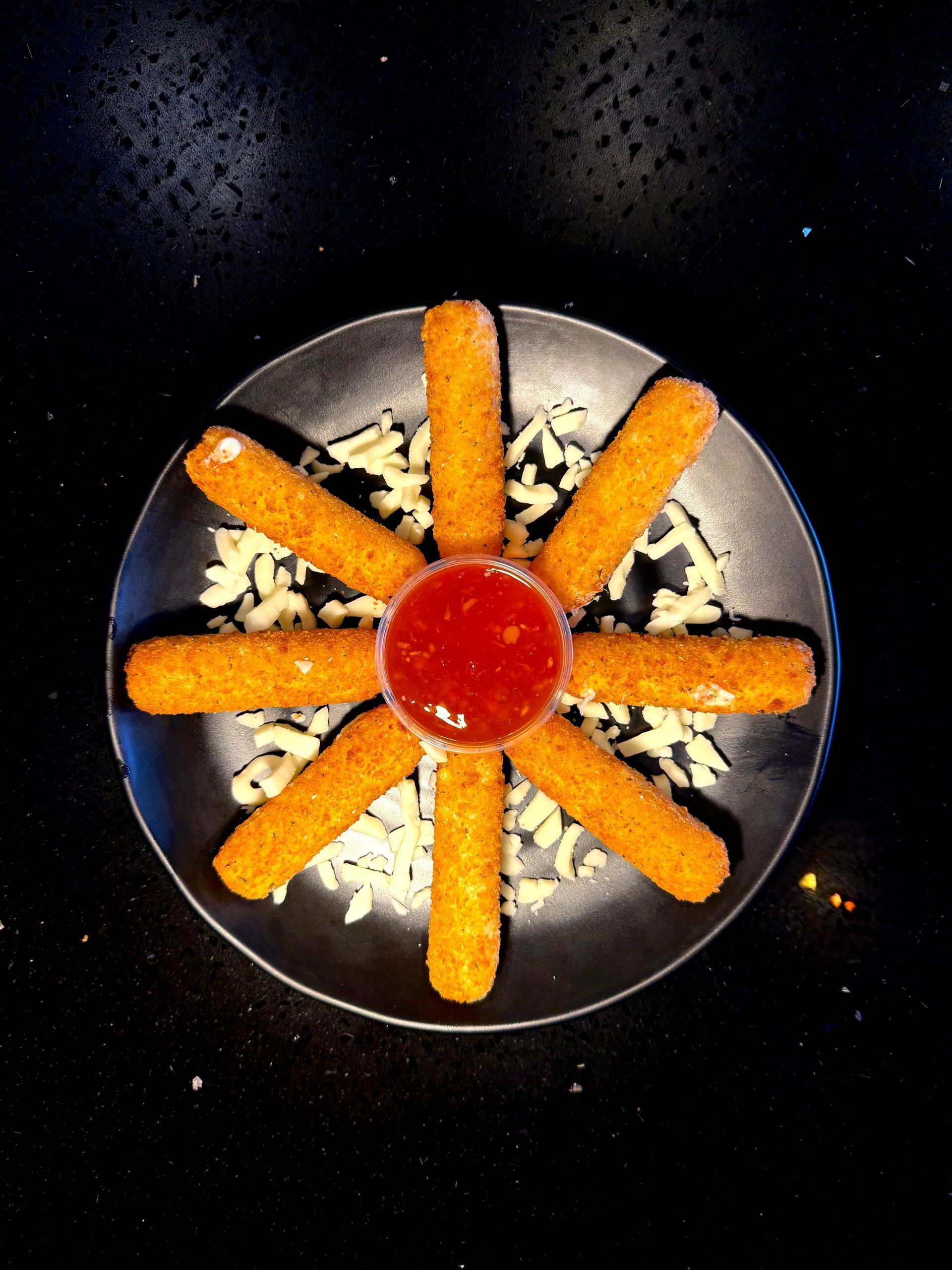 Mozzarella sticks arranged around a cup of marinara sauce on a bed of shredded cheese; black plate.