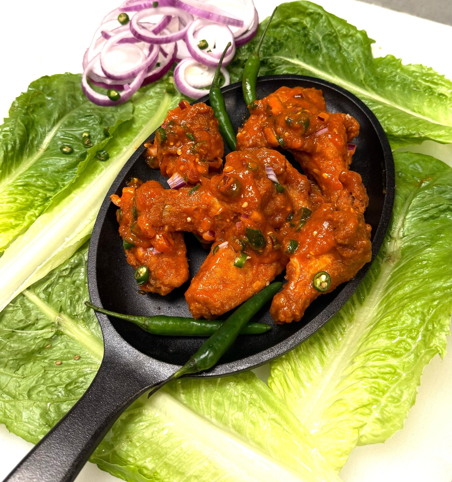 Spicy fried chicken wings on a black serving dish, surrounded by lettuce, red onion, and green chilies.