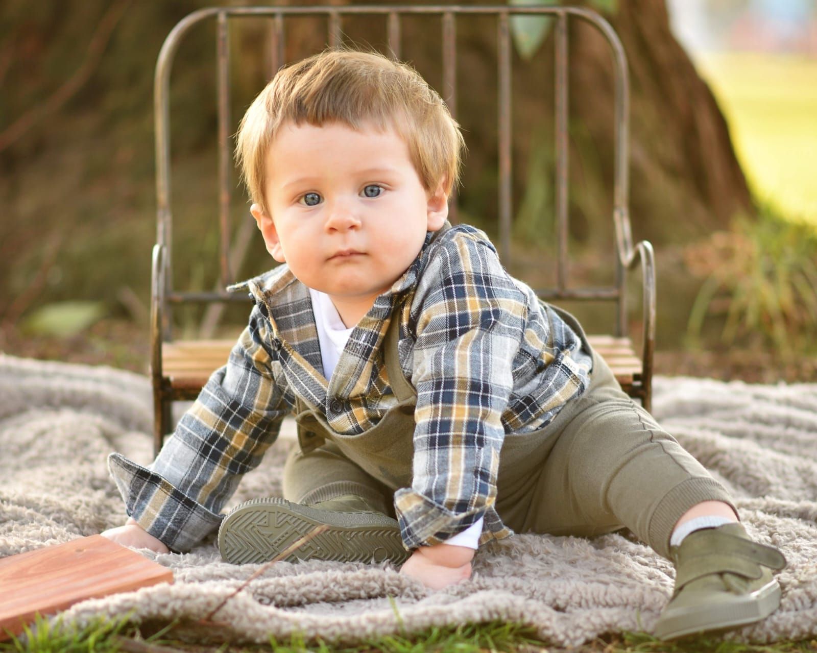 Bebé con camisa a cuadros y pantalones verdes gateando sobre una manta, con una pequeña silla de metal.