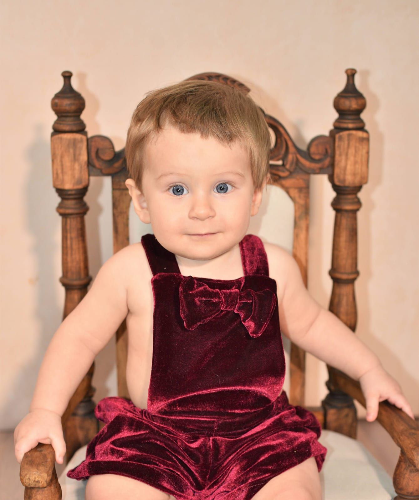 Bebé con un mono marrón con un lazo, sentado en una silla de madera.