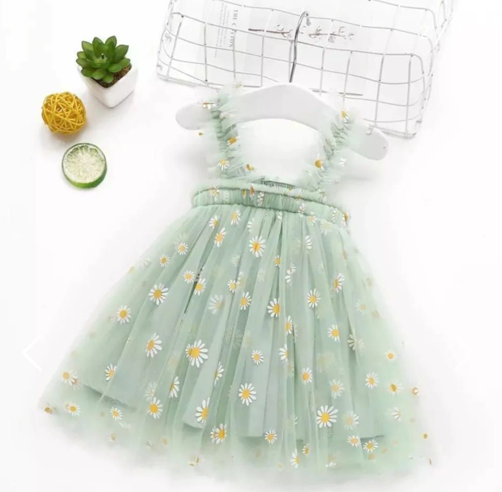 Vestido de tul con estampado de margaritas verdes en una percha blanca, junto a comida decorativa y una cesta de alambre.