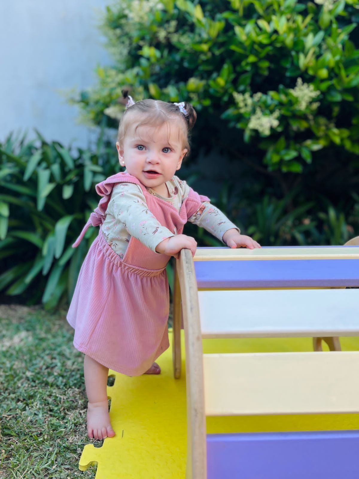 Niño con vestido rosa, apoyado en una estructura de madera, al aire libre en un día soleado.
