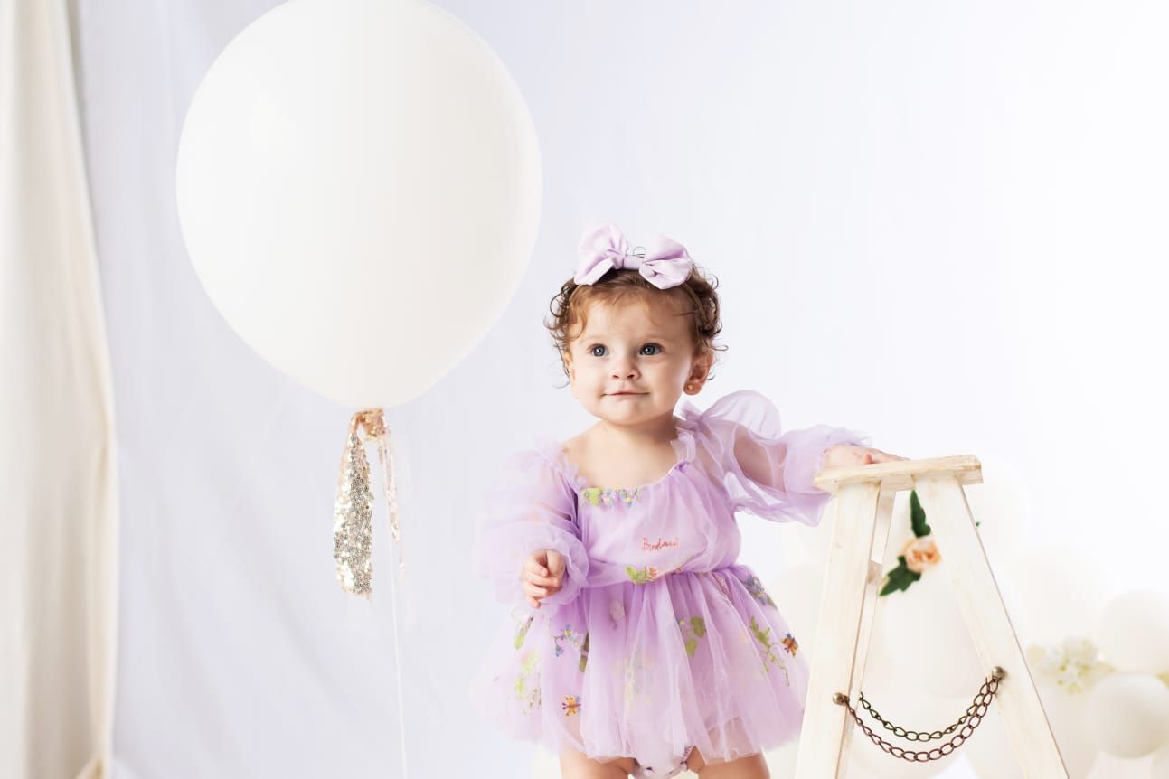 Un bebé con un vestido y una diadema color lavanda sonríe, sosteniendo un tipi decorado, cerca de un globo blanco.