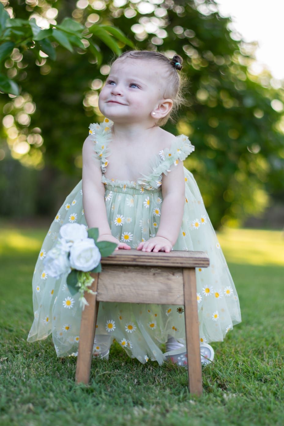 Niña con un vestido estampado de margaritas, de pie en un pequeño taburete de madera, mirando hacia arriba