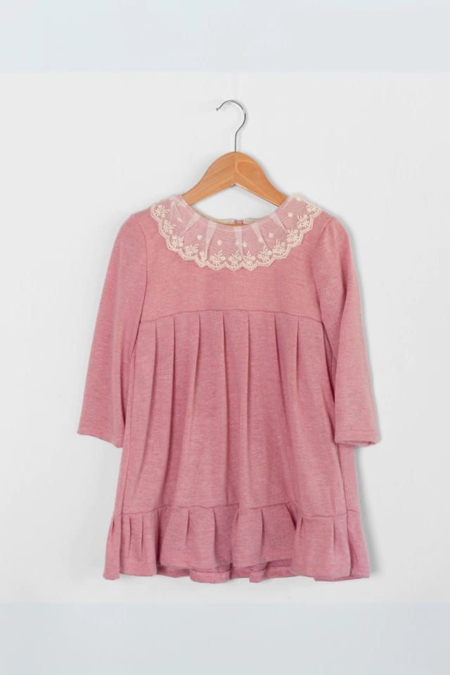 Vestido infantil de color rosa con cuello de encaje, mangas tres cuartos y bajo con volante, colgado en una percha de madera.