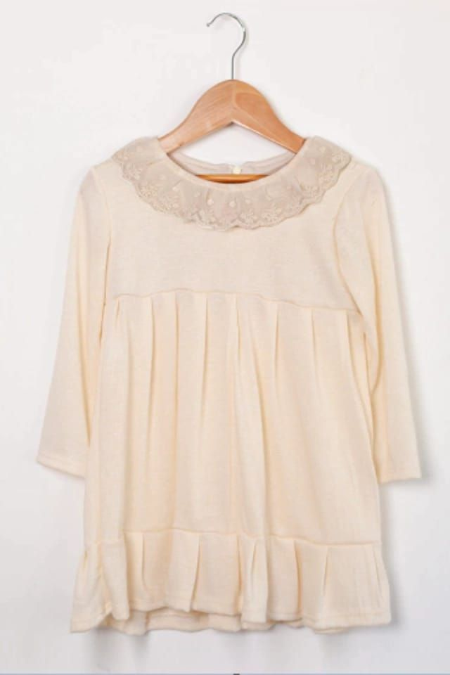 Vestido infantil de color rosa con cuello de encaje, mangas tres cuartos y bajo con volante, colgado en una percha de madera.