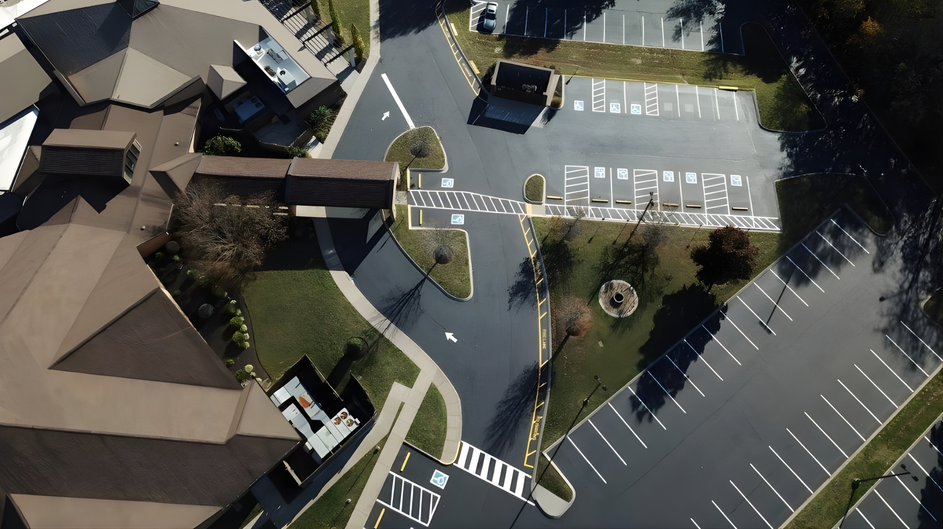 Aerial view of a white, L-shaped building with surrounding black asphalt parking and road, nestled in green trees and grass.